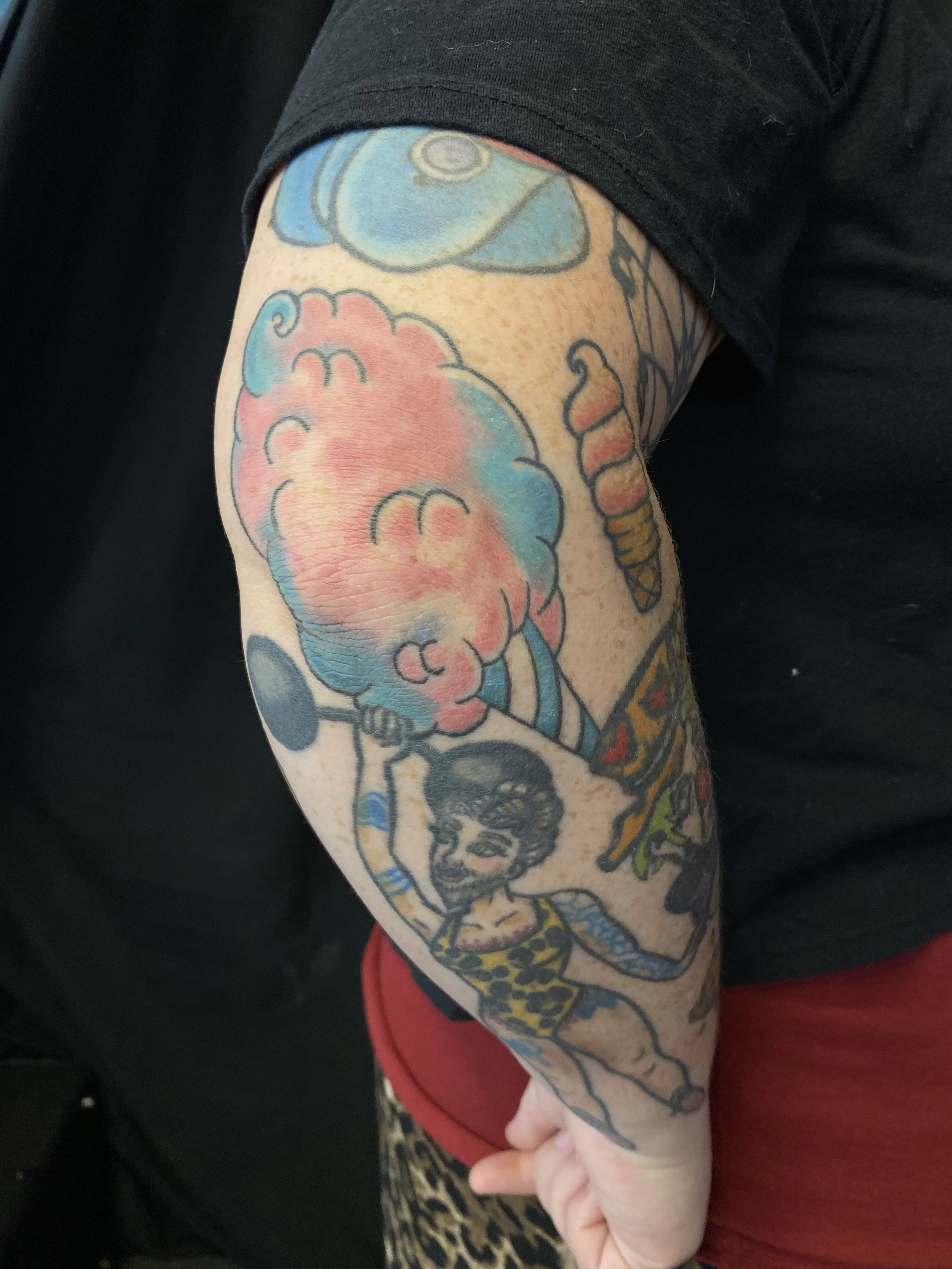 Colorful tattoo sleeve on a person's arm featuring a cartoon-style character with a large head, smiling face, and vibrant colors including pink, blue, and yellow.