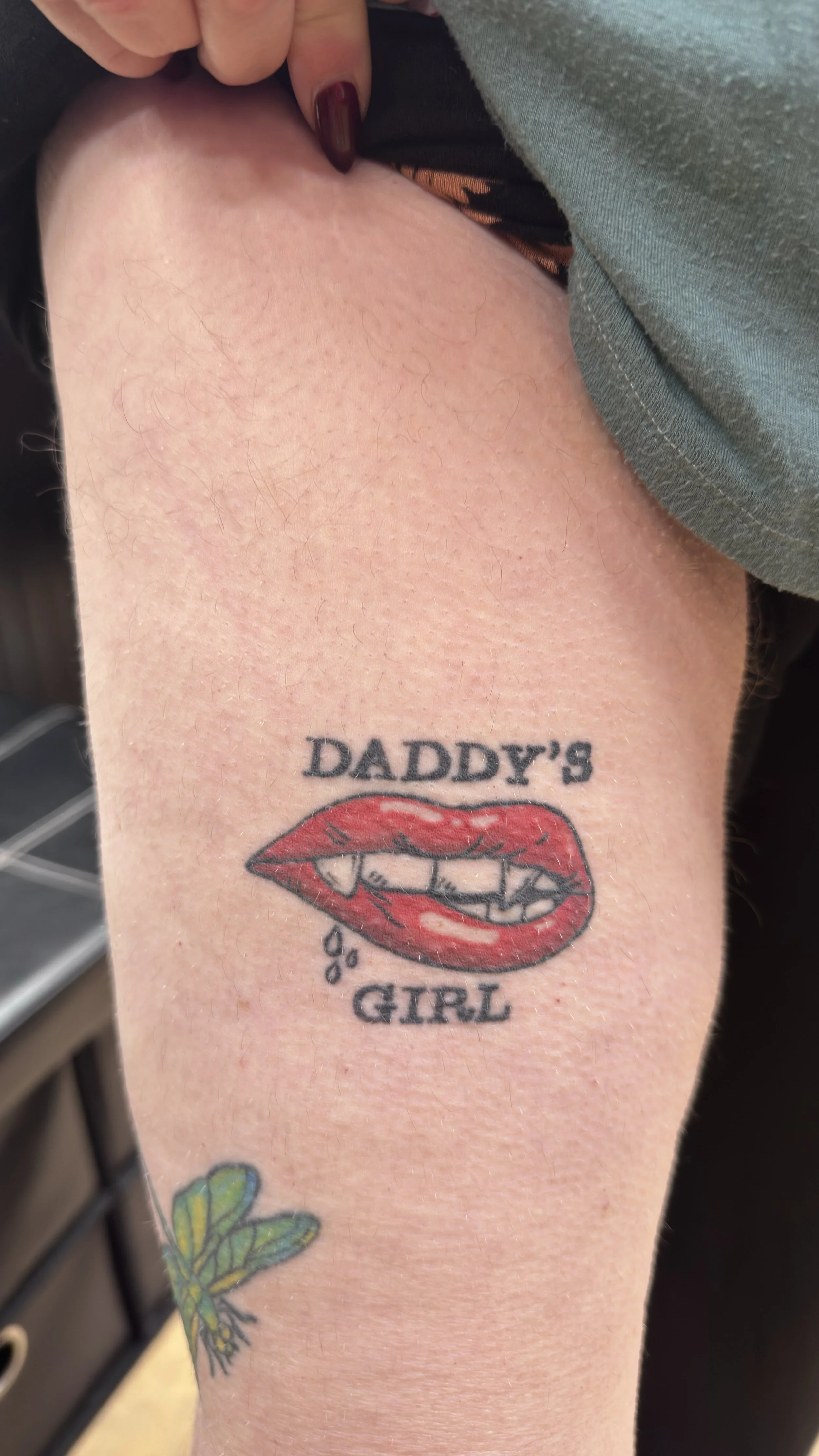 Tattoo of a pair of lips with the words "Daddy's Girl" above and below it, on a person's arm.