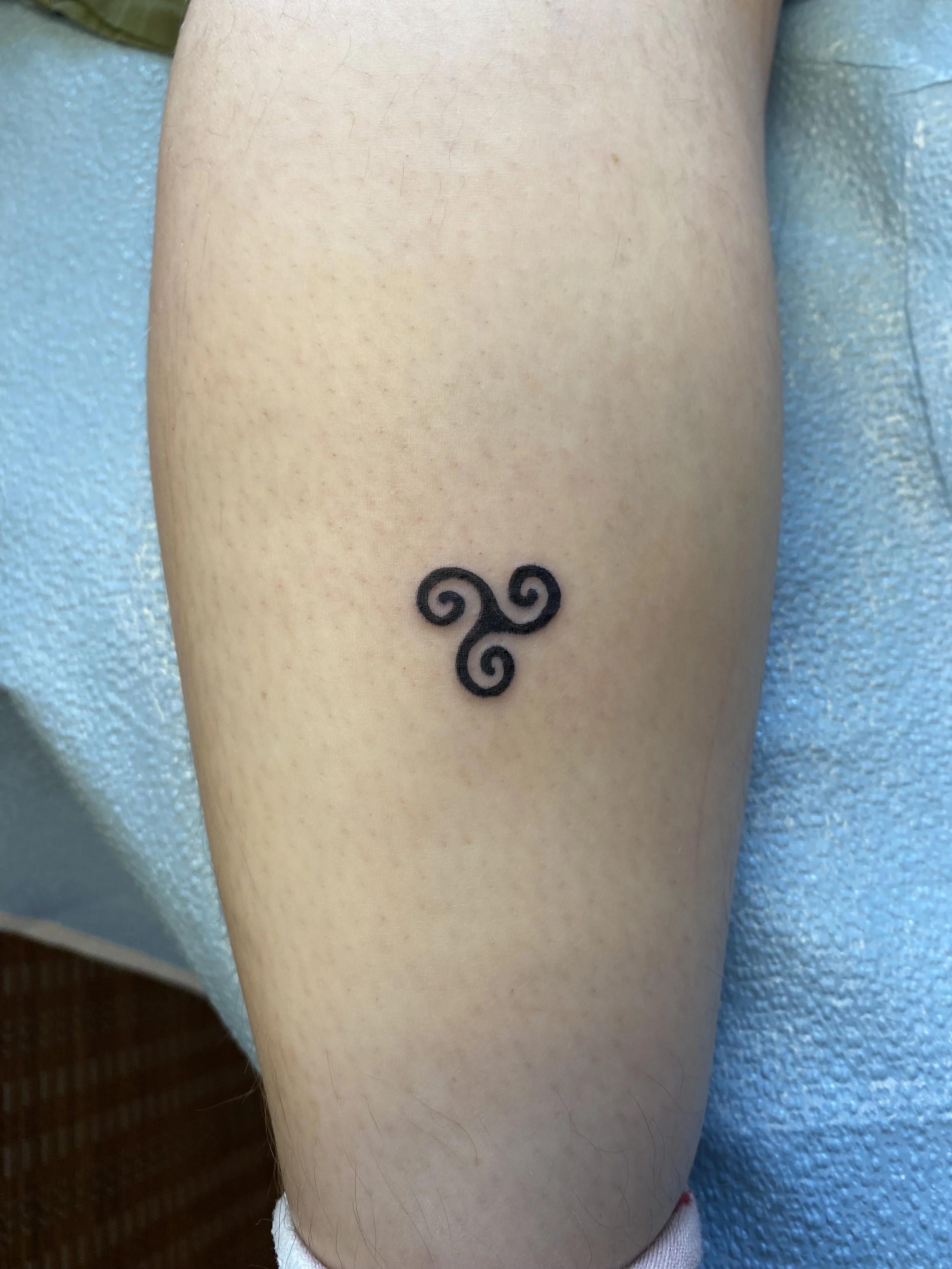 Close-up of a person's arm with a small black tattoo of a triskelion symbol.