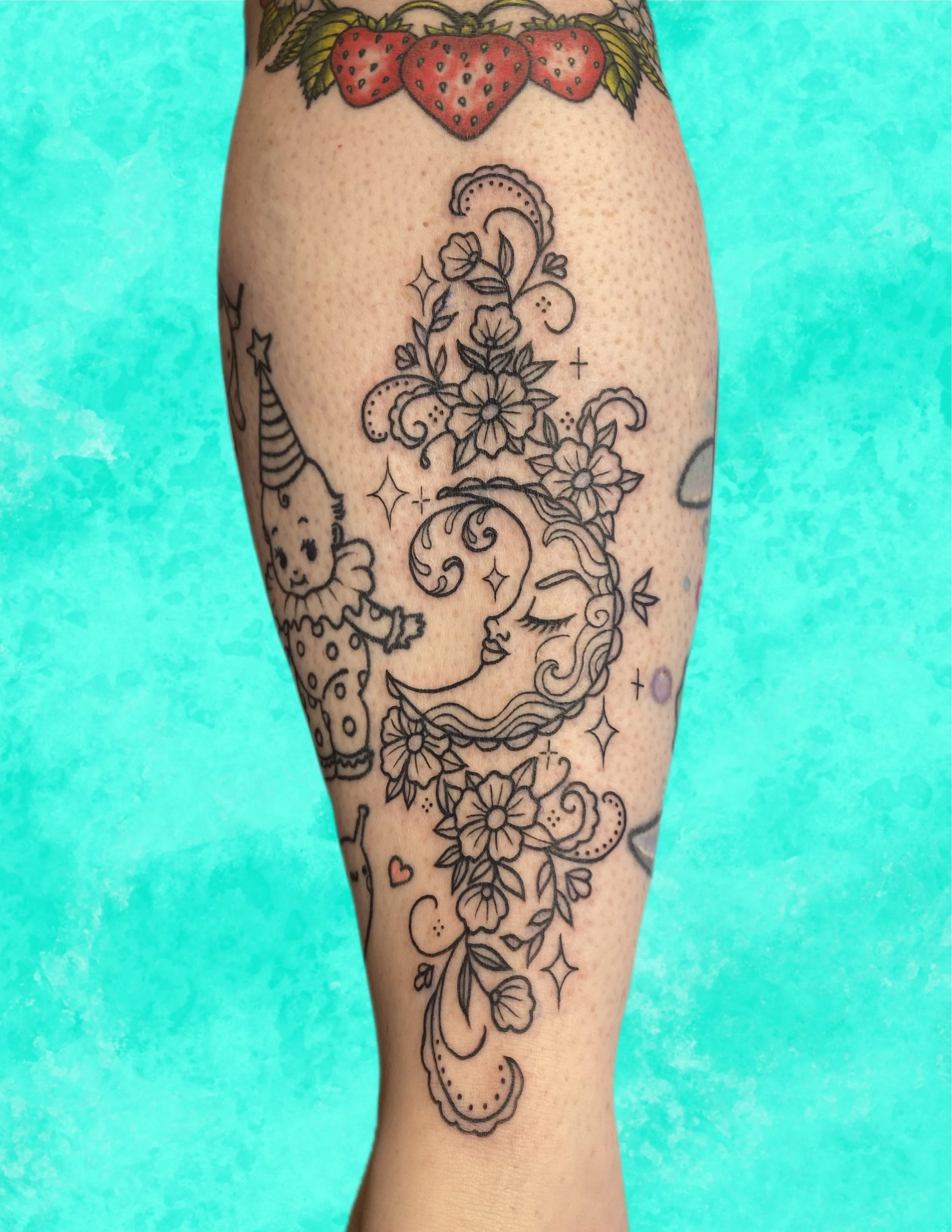 Tattoo of flowers and decorative patterns on a person's leg, with a strawberry-themed tattoo at the top and various floral and whimsical designs down the leg, set against a turquoise background.