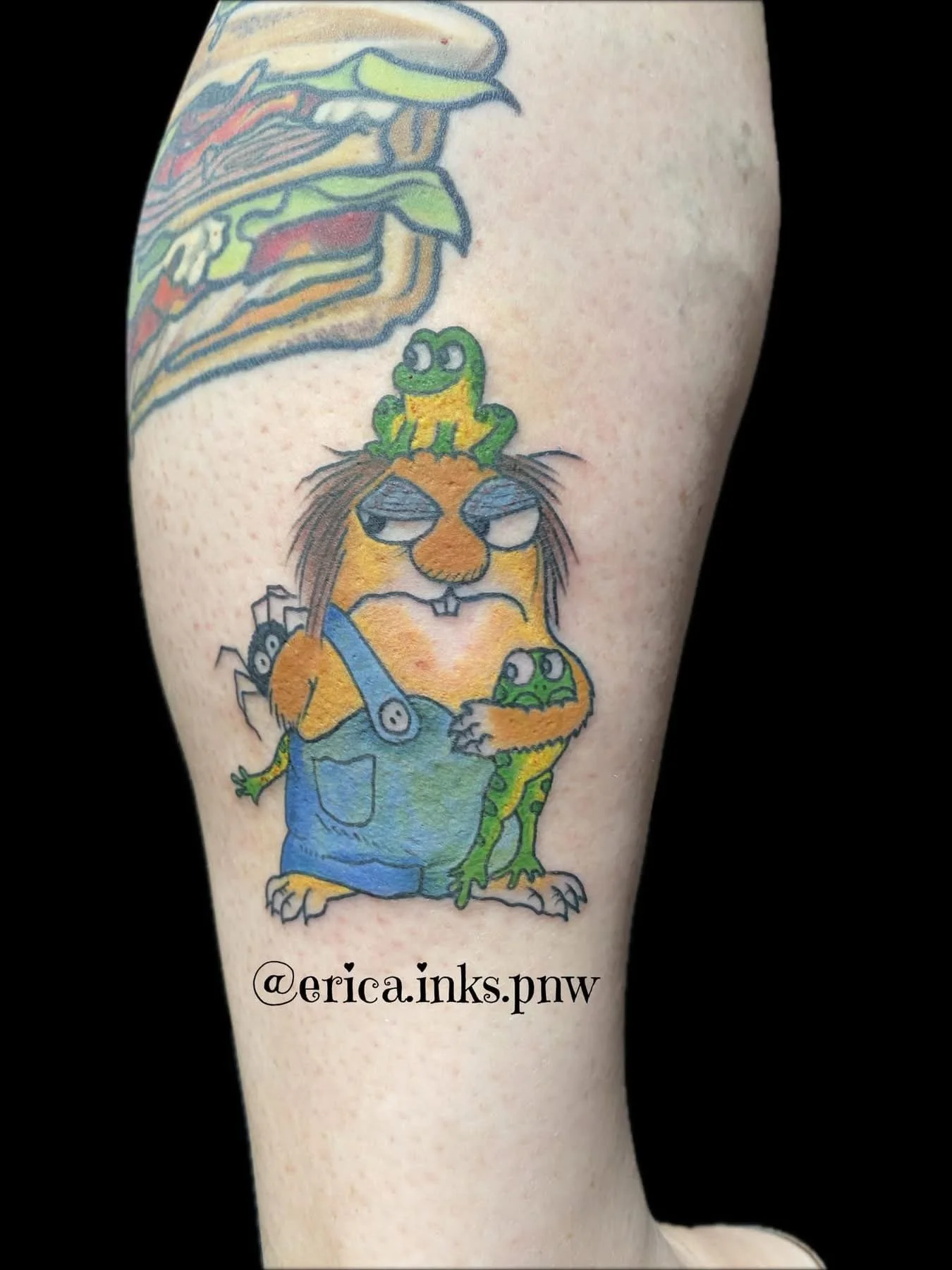 Colorful tattoo of a lion wearing overalls, with a frog on its head, a frog in its hand, and spider on its back, on a person's arm.