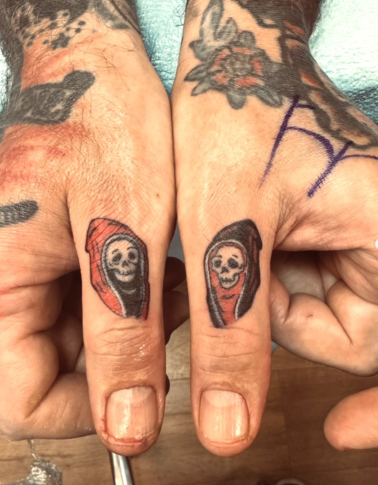 Close-up photo of two fingers with matching tattoos of smiling skulls in red and black ink, with additional tattoos on the backs of the hands and around the fingers.