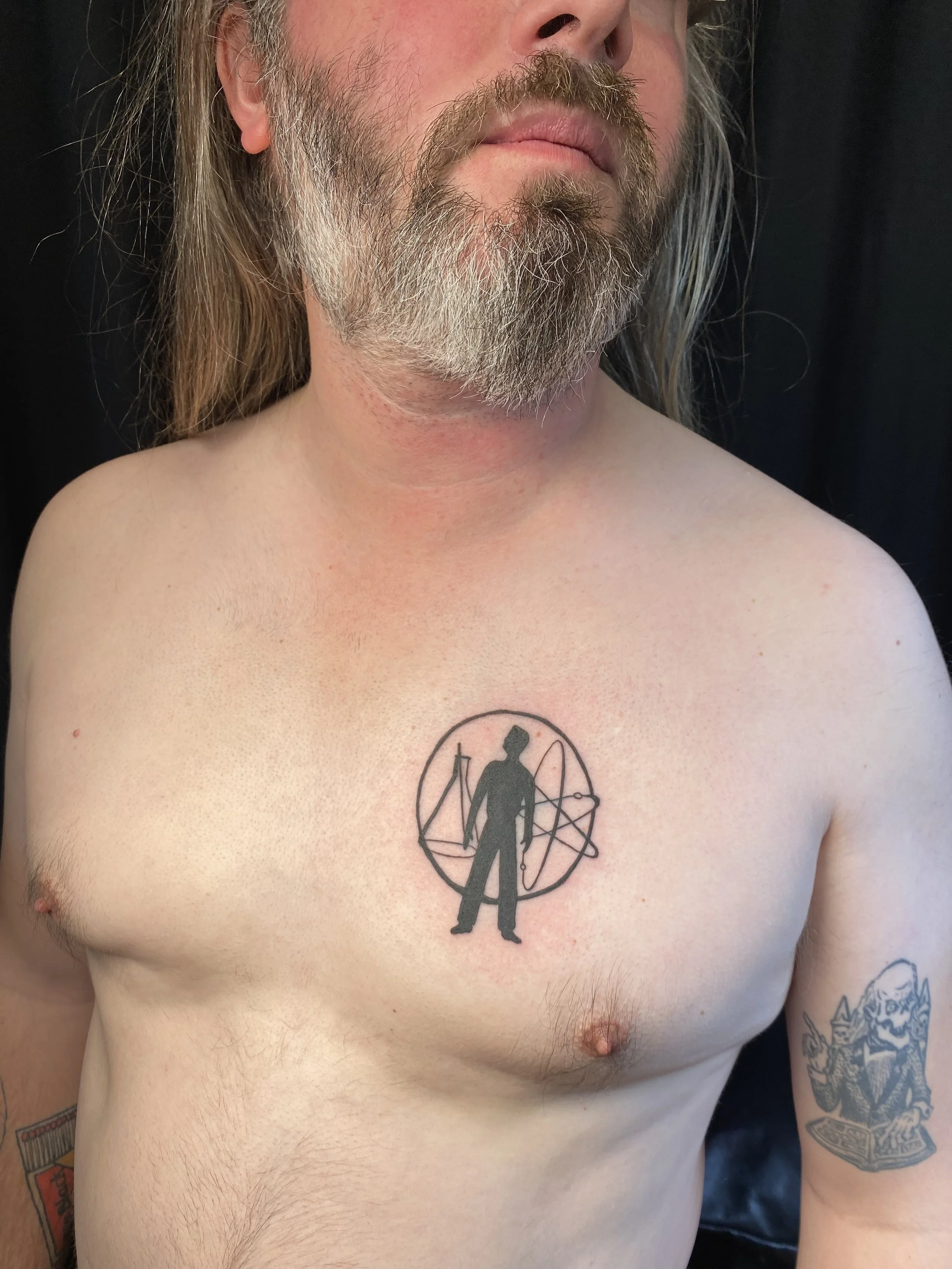 A shirtless man with long hair and a beard, showing a tattoo of a liberty statue with a circle and geometric elements on his chest.