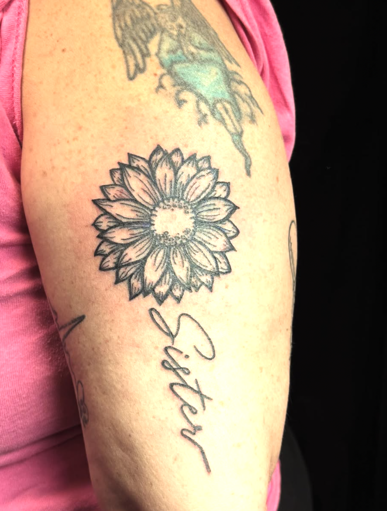 A close-up of a arm with a sunflower tattoo, black outline, and some other tattoos including a bird and a word or phrase in cursive. The person is wearing a pink shirt or top.