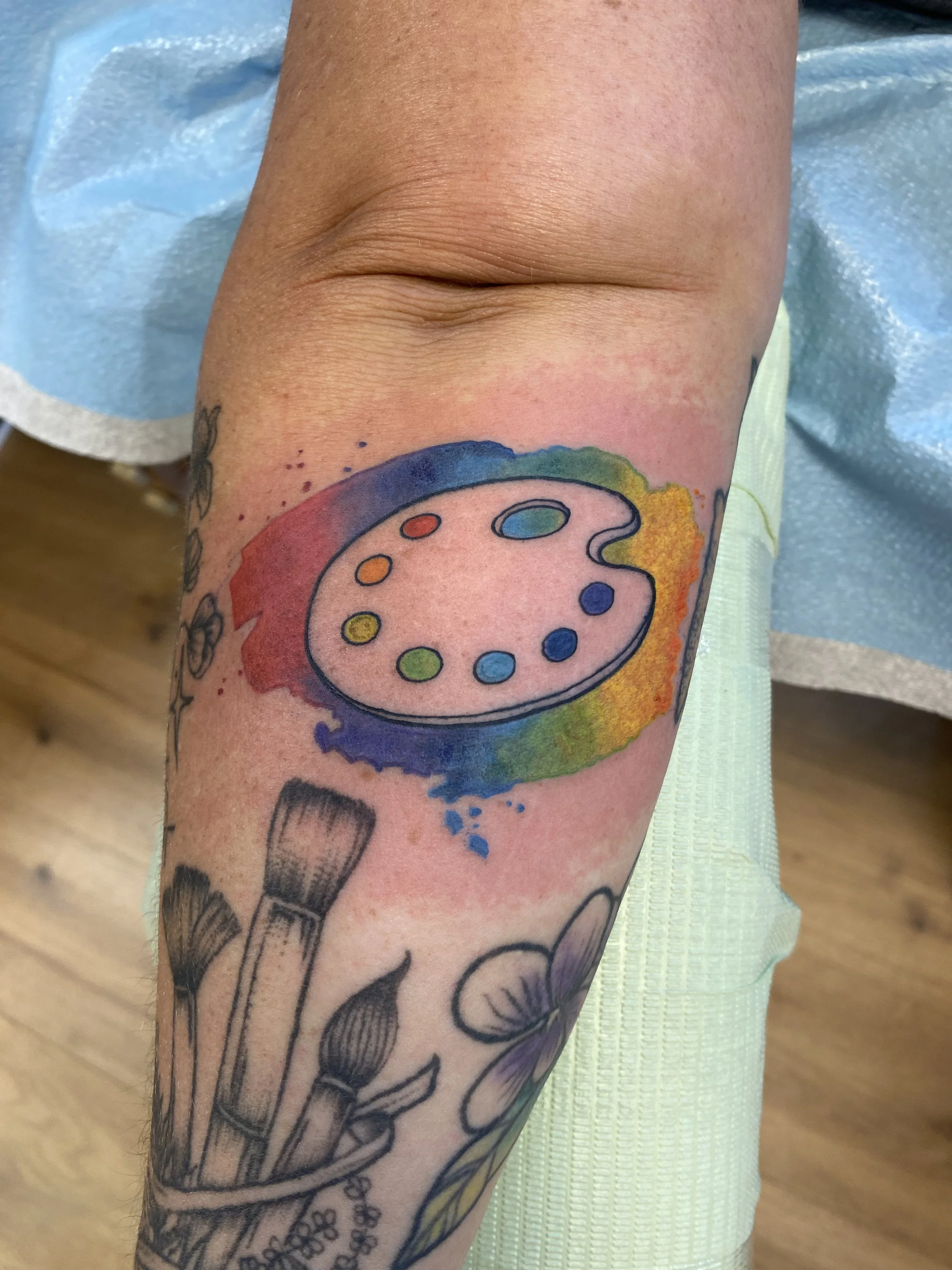 Close-up of a person's arm with a colorful tattoo featuring a rainbow palette and a paint palette design. Several other tattoos are visible including flowers and abstract drawings.