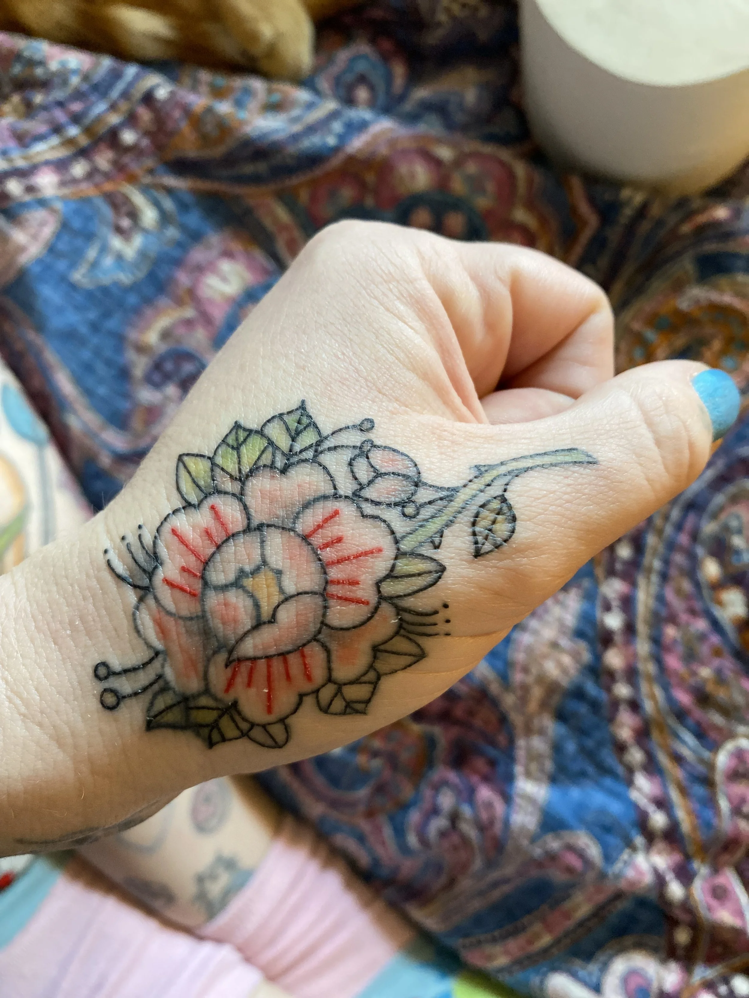A person's hand with a tattoo of a flower, featuring red, yellow, and green ink, resting on a colorful, patterned fabric.