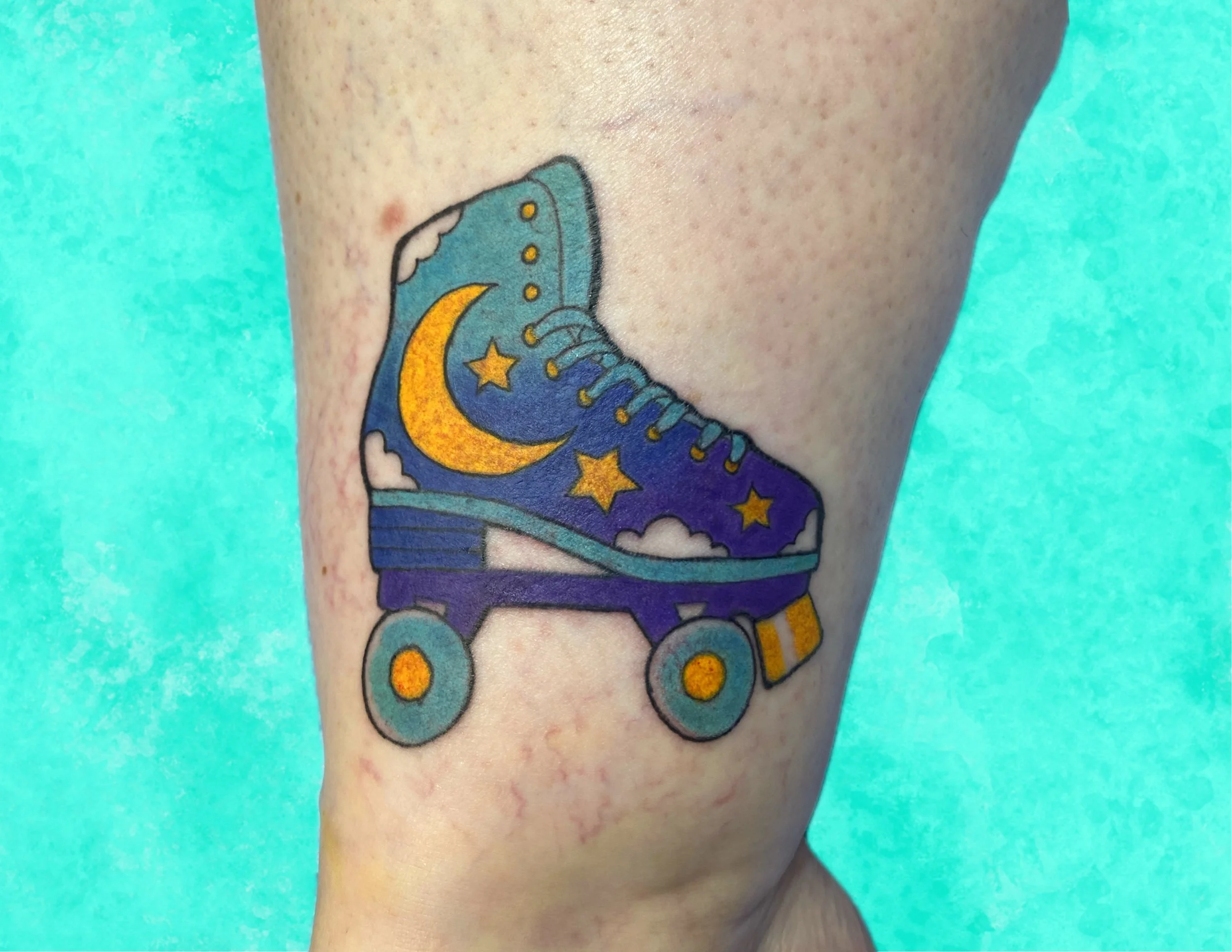Colorful tattoo of a space-themed sneakers with a yellow crescent moon and yellow stars, on a person's arm against a turquoise background.
