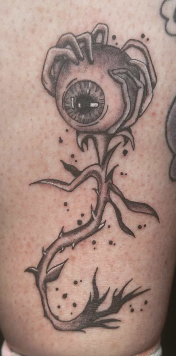 Tattoo of a flower with a human eye at the center, with thorny stem and leaves.