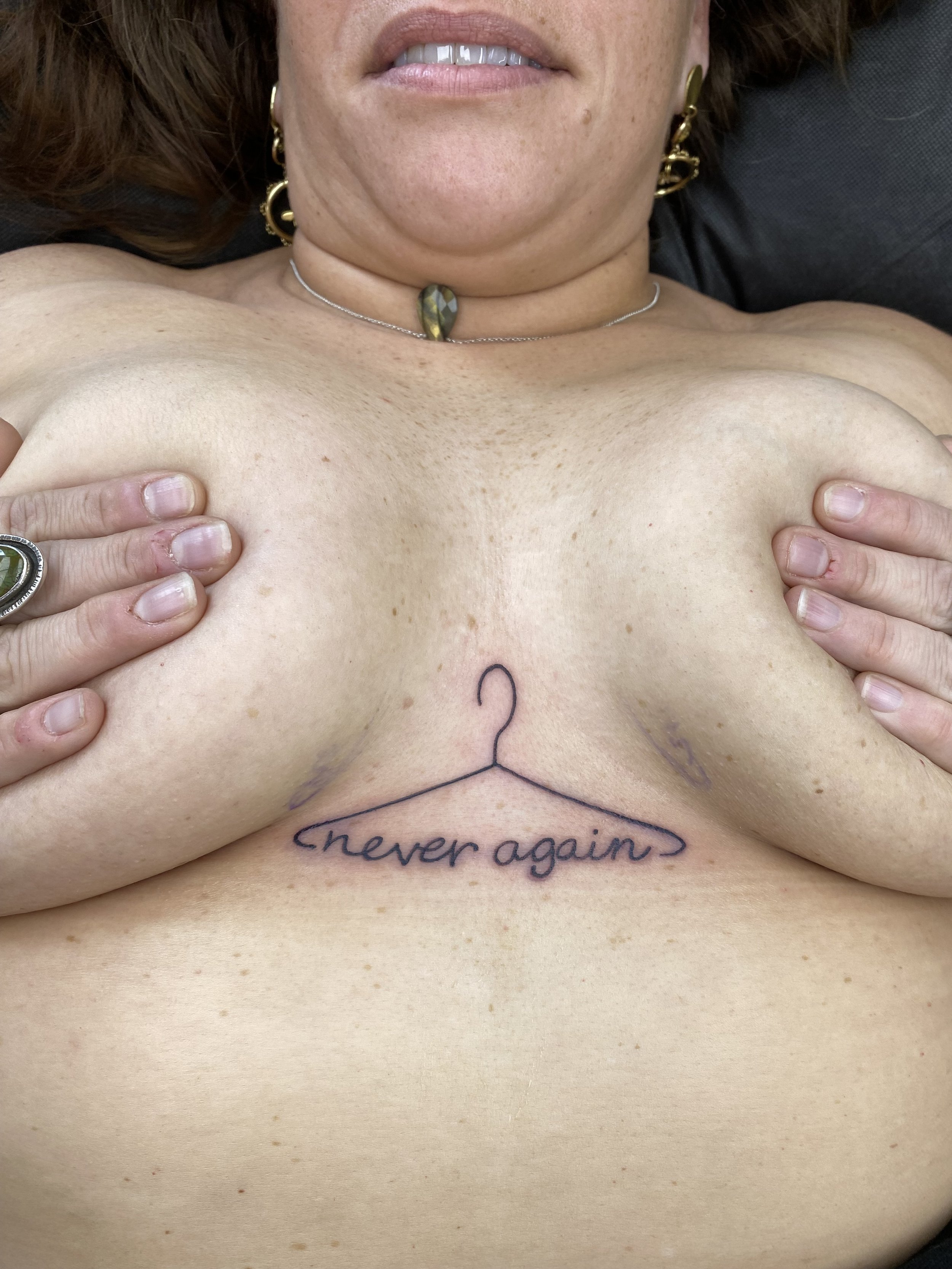 A close-up of a woman's upper body showing her bare chest, hands holding her breasts, a tattoo that says 'never again' below her chest, and a necklace with a green stone. She is wearing earrings and has a mole on her chin.