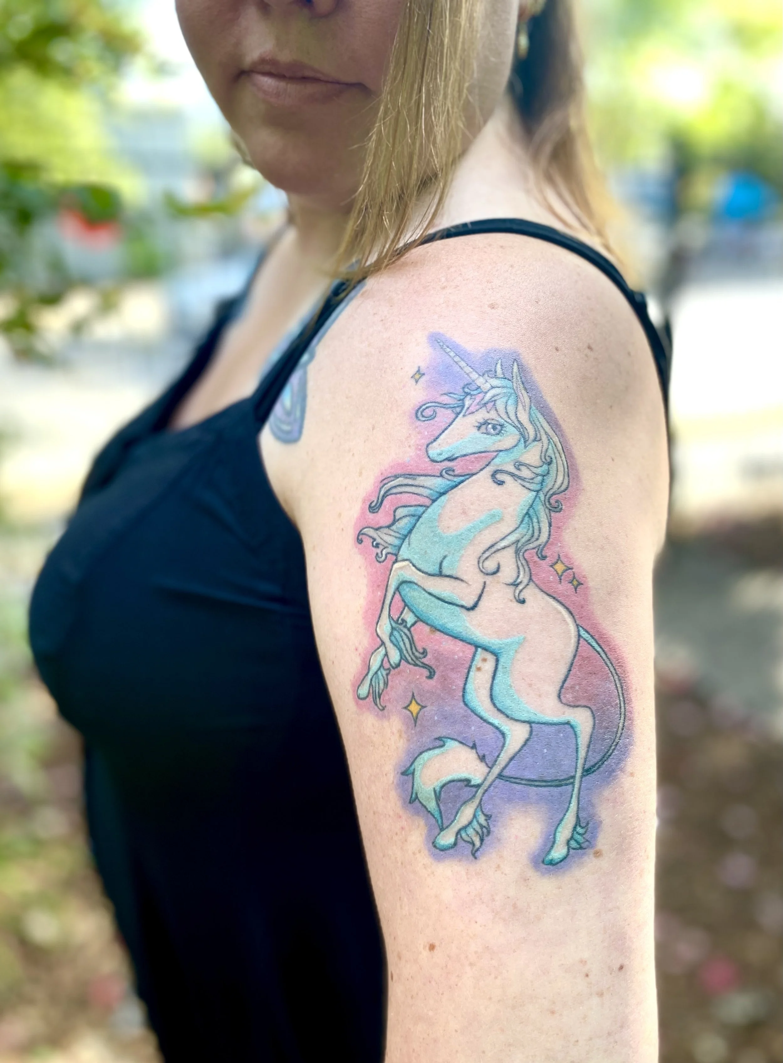 Close-up of a woman with a colorful unicorn tattoo on her upper arm, wearing a black sleeveless top, outdoors with blurred greenery in the background.