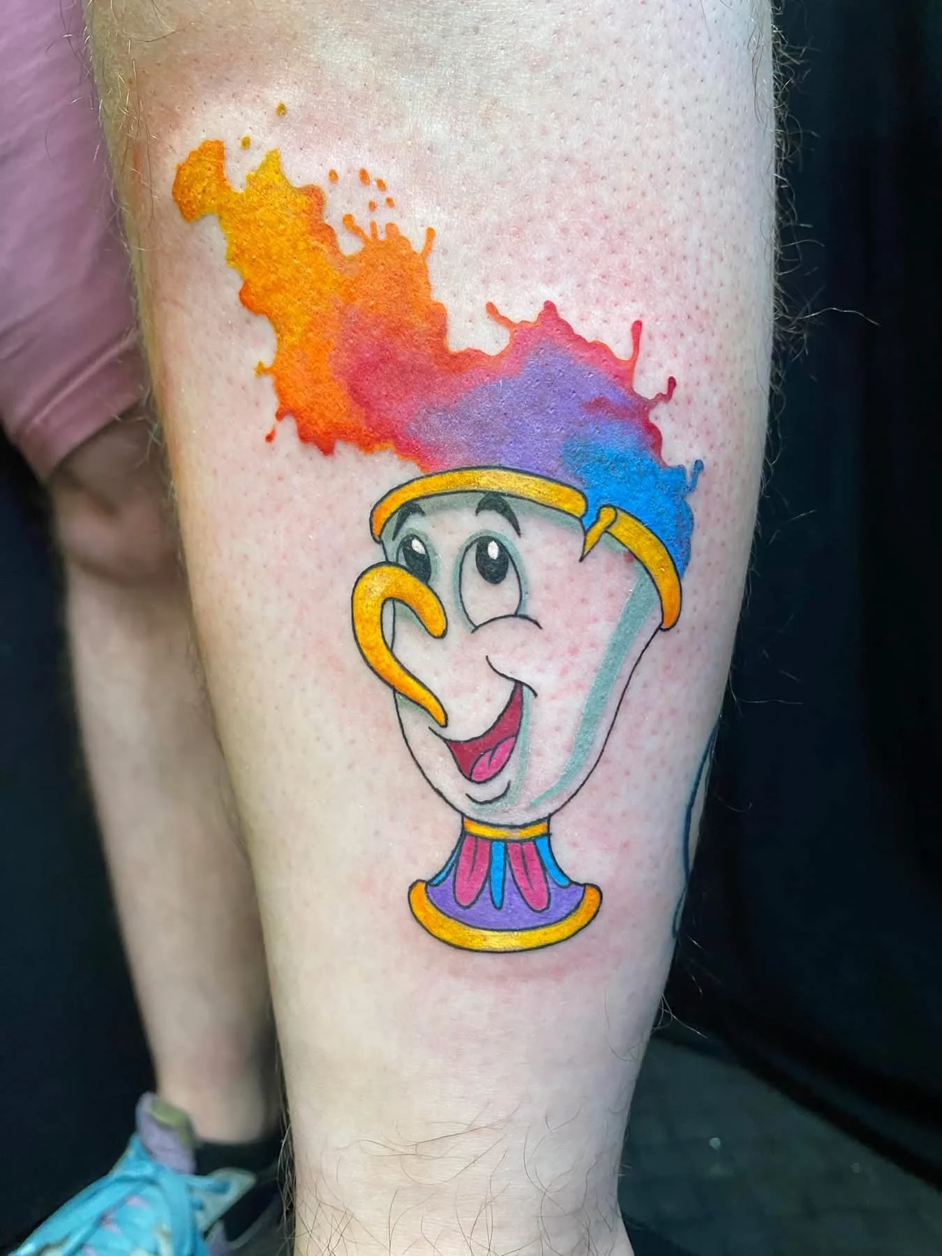 Tattoo of a cartoon clown with a big smile, holding a colorful splash of watercolor paint overhead.