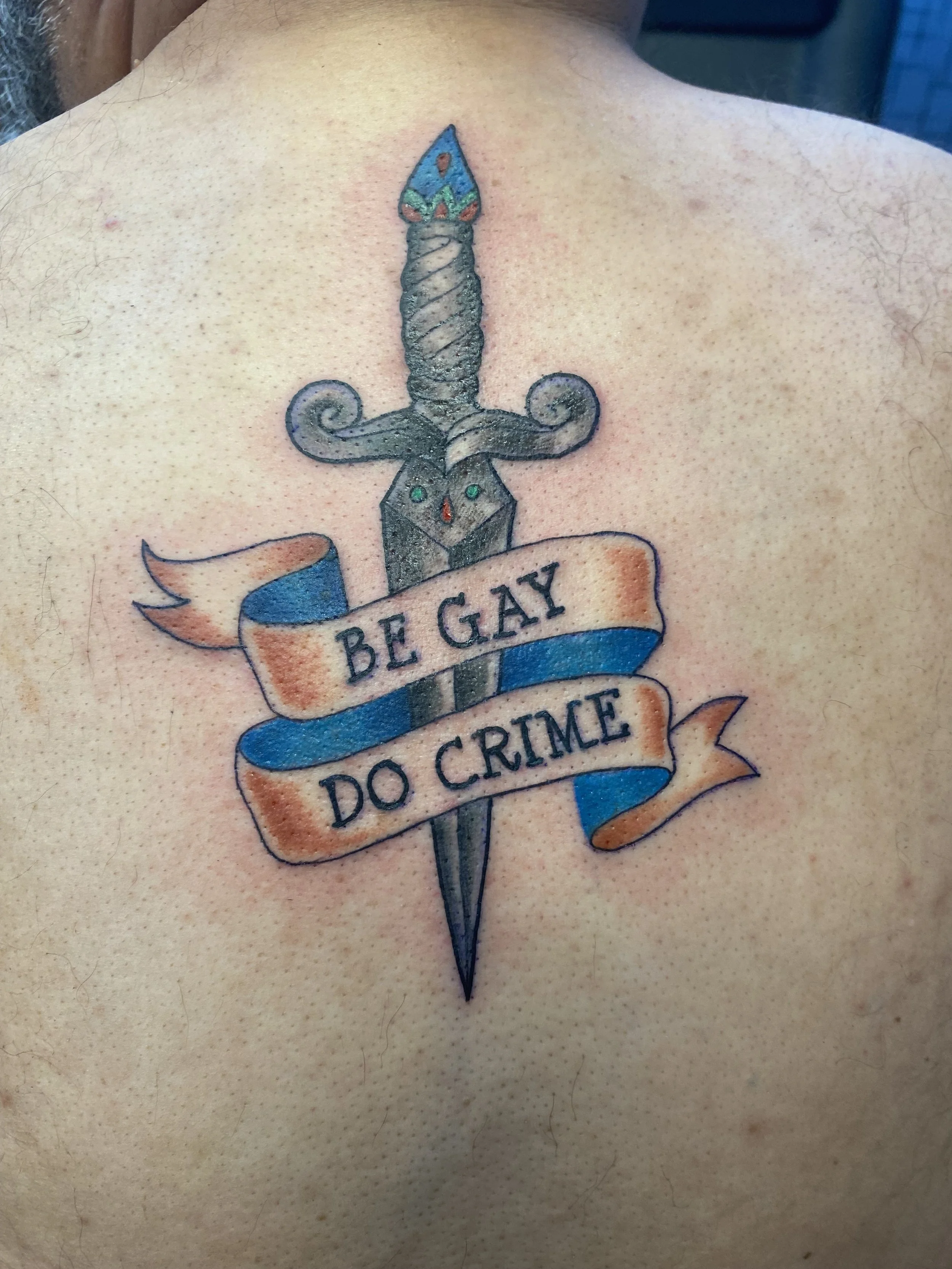 Close-up of a tattoo on a person's chest depicting a dagger with a colorful handle and a banner that reads 'BE GAY DO CRIME'.