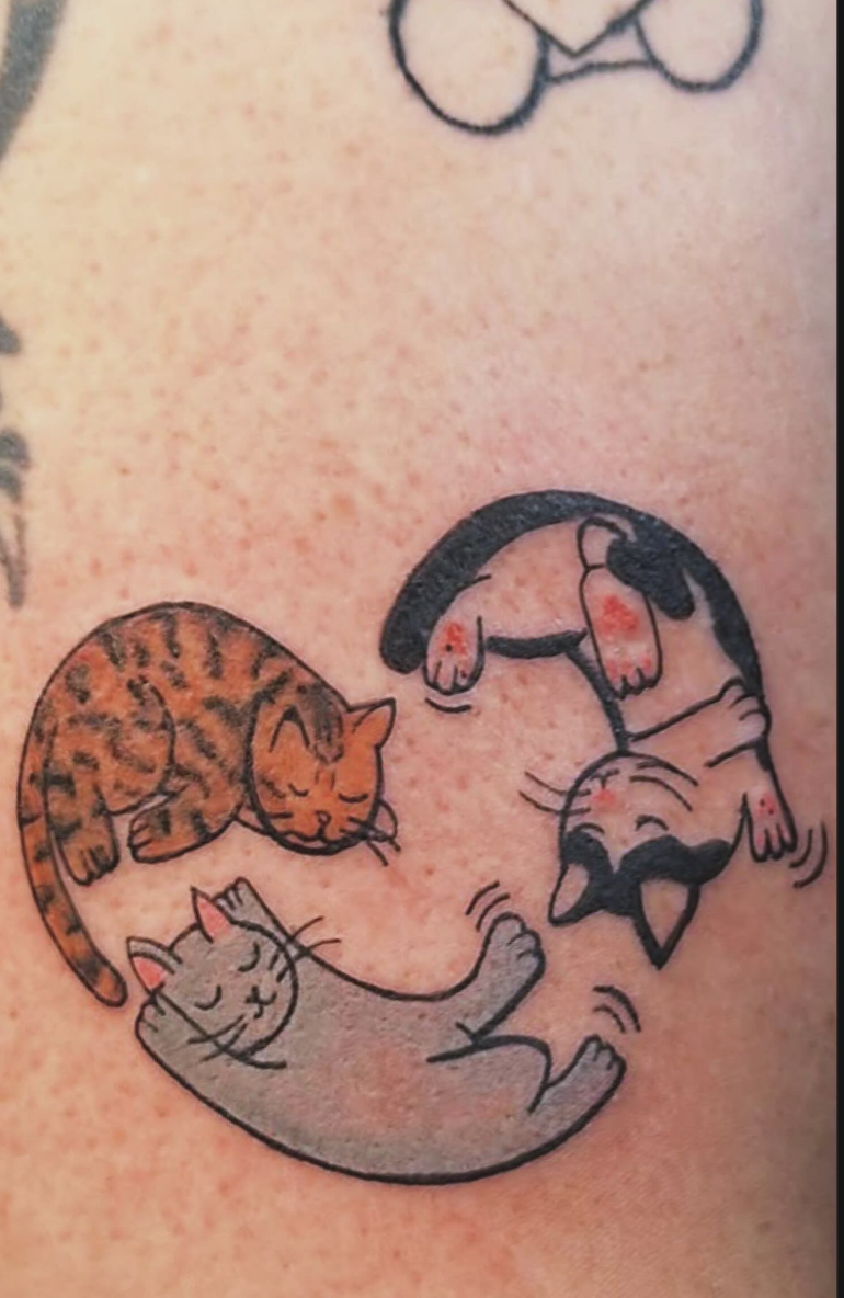 Three whimsical cat tattoos: one with orange and black stripes, one gray with pink accents, and one black and white, all sleeping or curled up, on a person's skin.