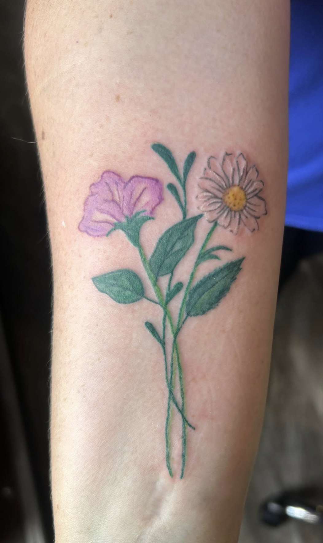 A tattoo of two flowers, one purple and one yellow, with green leaves on a person's arm.