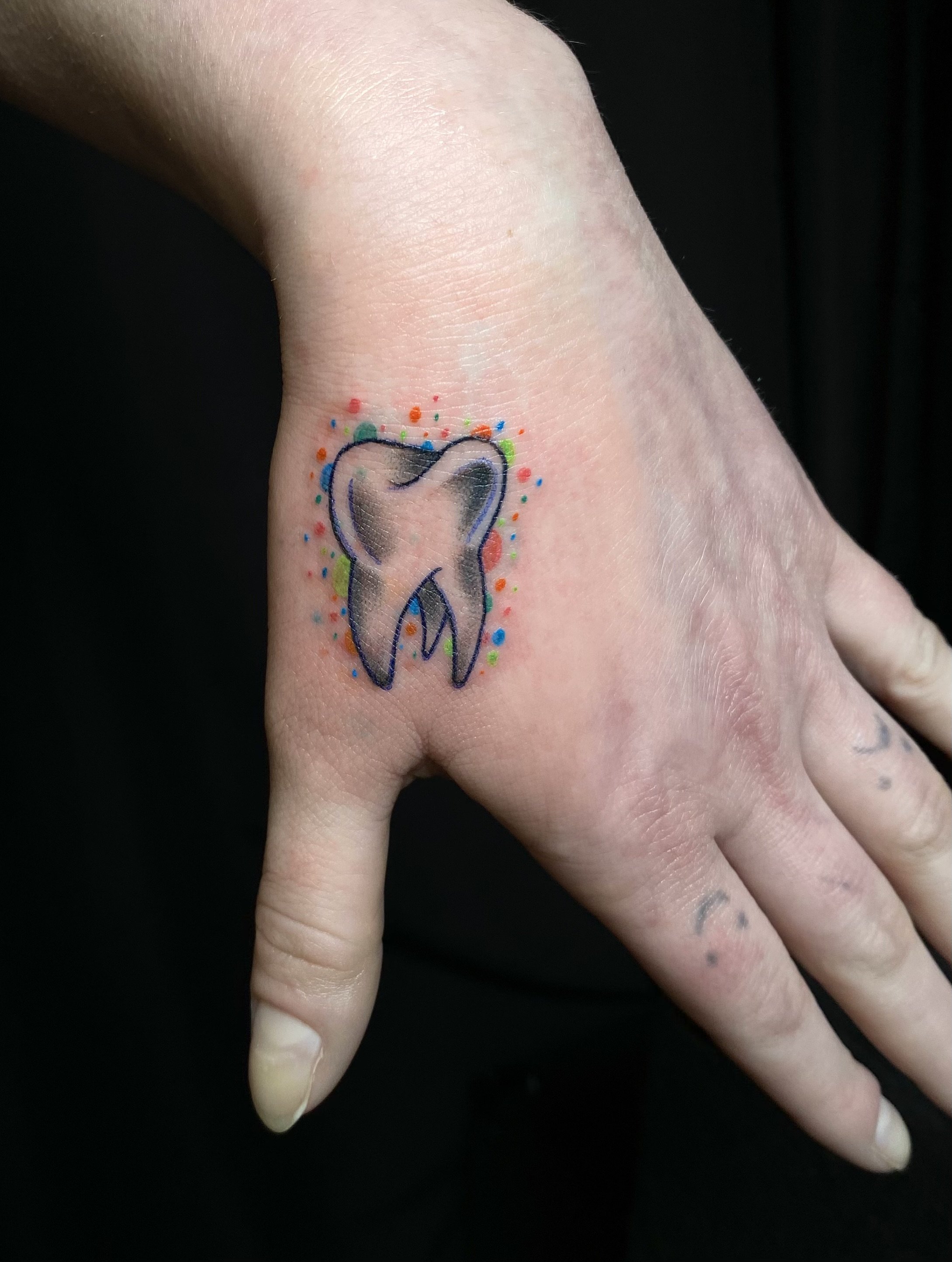 A hand with a tattoo of a tooth on the finger, surrounded by colorful dots on the skin, against a black background.