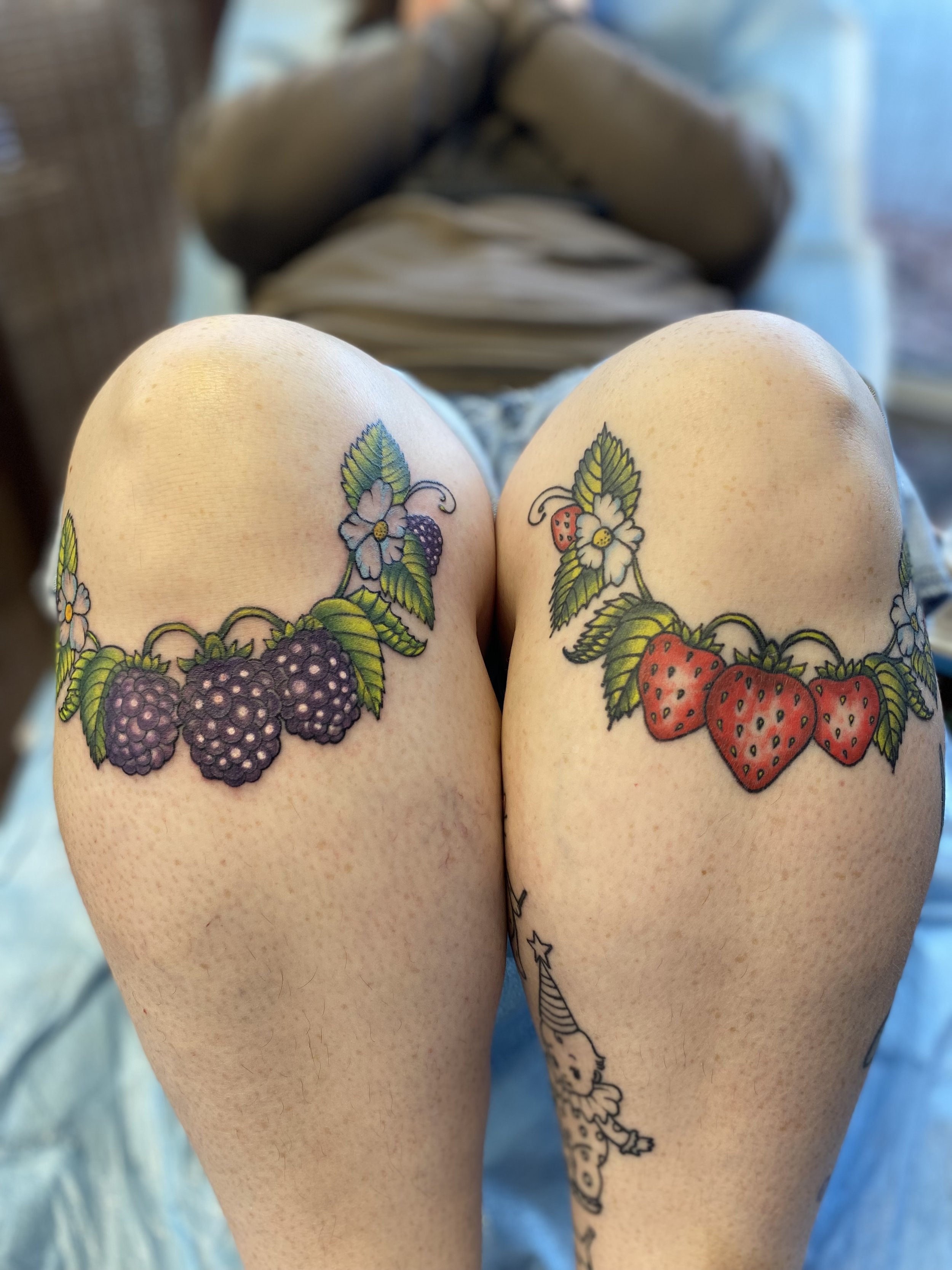 Close-up of a person's thighs with colorful tattoos of blackberries and strawberries, along with floral and leafy designs, on their upper legs.