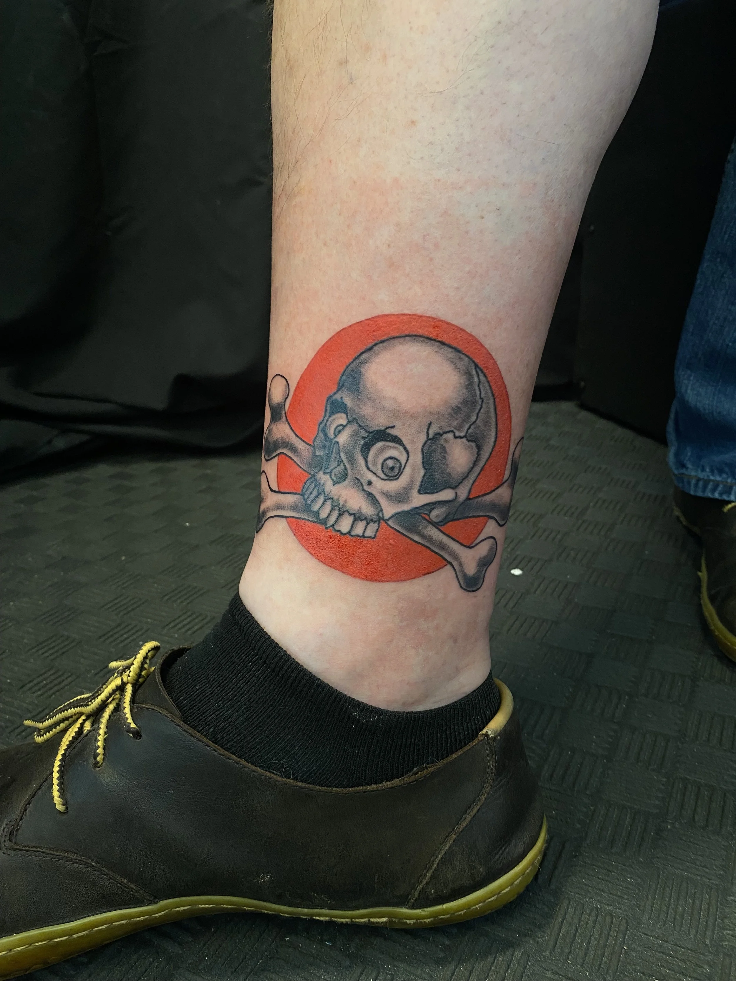 Tattoo of a skull with a bone across the top, within a red circle, on a person's lower leg.