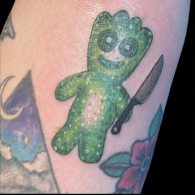 Tattoo of a green plush toy with big eyes and a big smile, holding a knife.