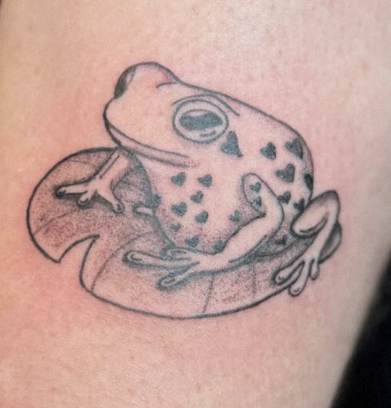 Black and gray tattoo of a frog sitting on a lily pad, with detailed skin and closed eyes.