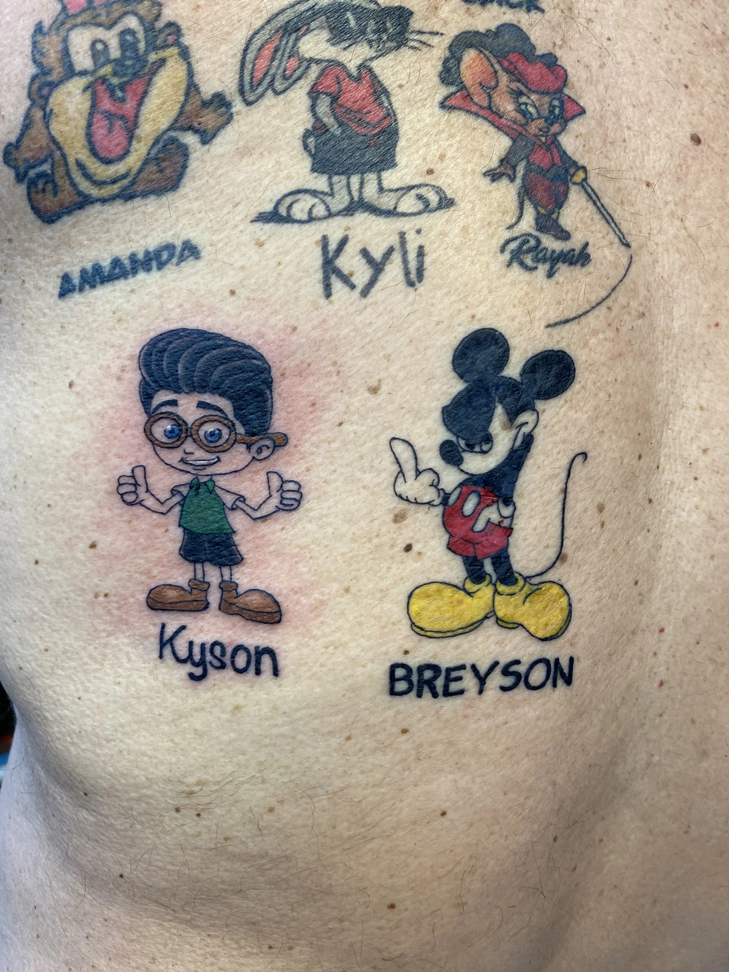 Close-up of a tattoo featuring cartoon characters and names, including Homer Simpson labeled "Amora," Kyli, Mickey Mouse labeled "Reyash," and Mickey Mouse labeled "Kyson" and "Breyon."