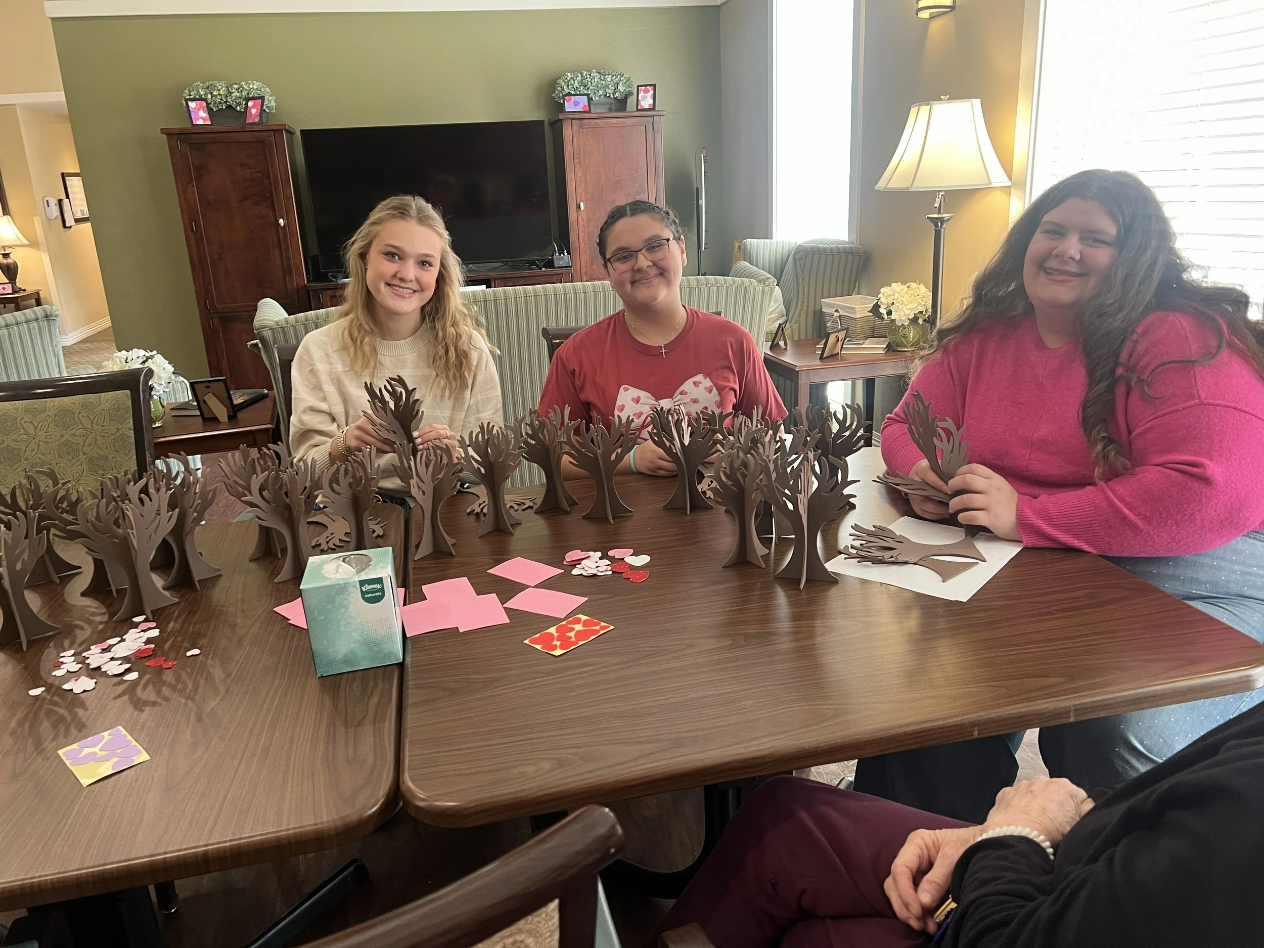Three young women smiling at a table with paper reindeer crafts, Valentine's Day decorations, and a tissue box, seated in a cozy, well-lit room with green walls, a large TV, and framed photos.