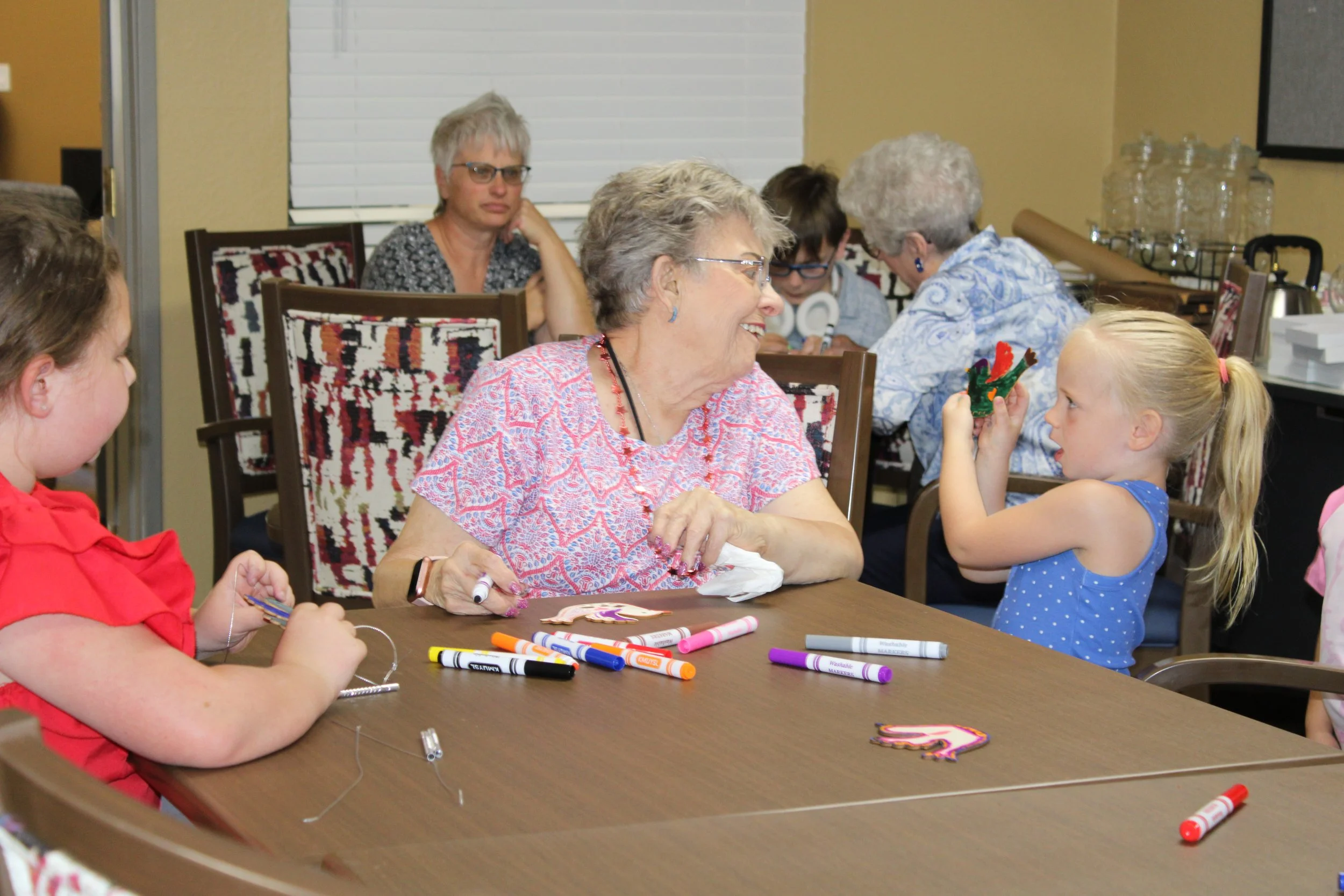 Elderly woman smiling and talking to a young girl with blond hair, wearing a blue dress, handing her a small craft item in a room with several other people at a table, markers, and decorated cutouts.