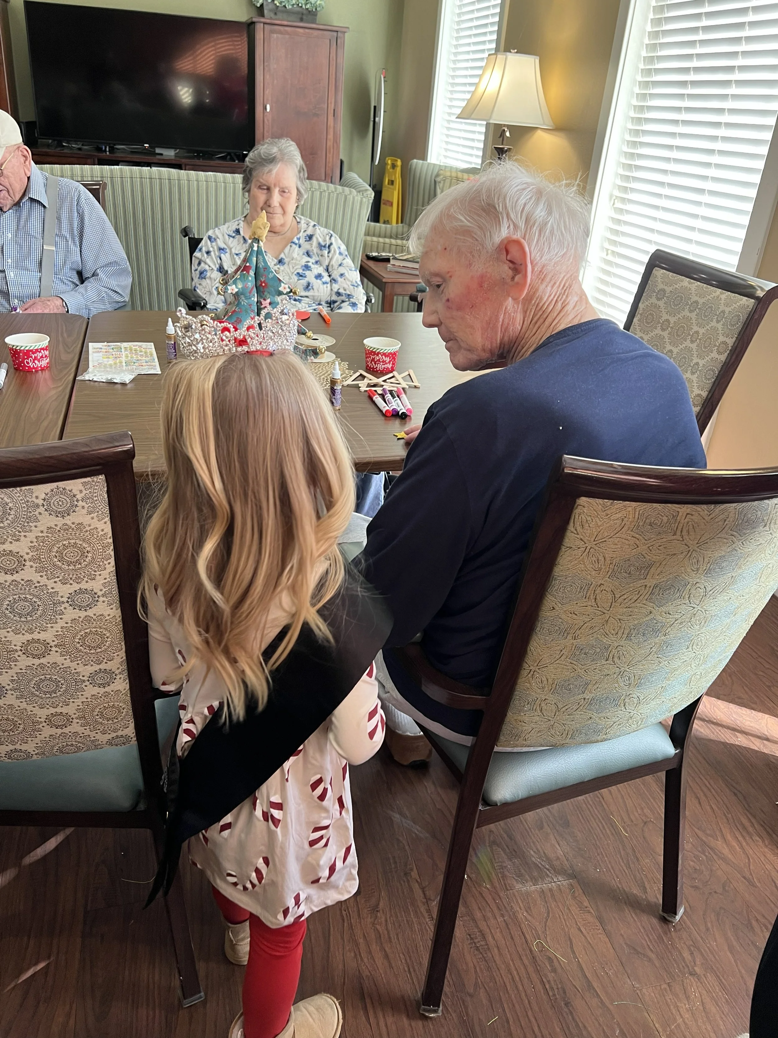 A group of elderly people and a young girl gathered around a dining table decorated for Christmas, with a small decorated tree and holiday cups, in a brightly lit room with windows and blinds.