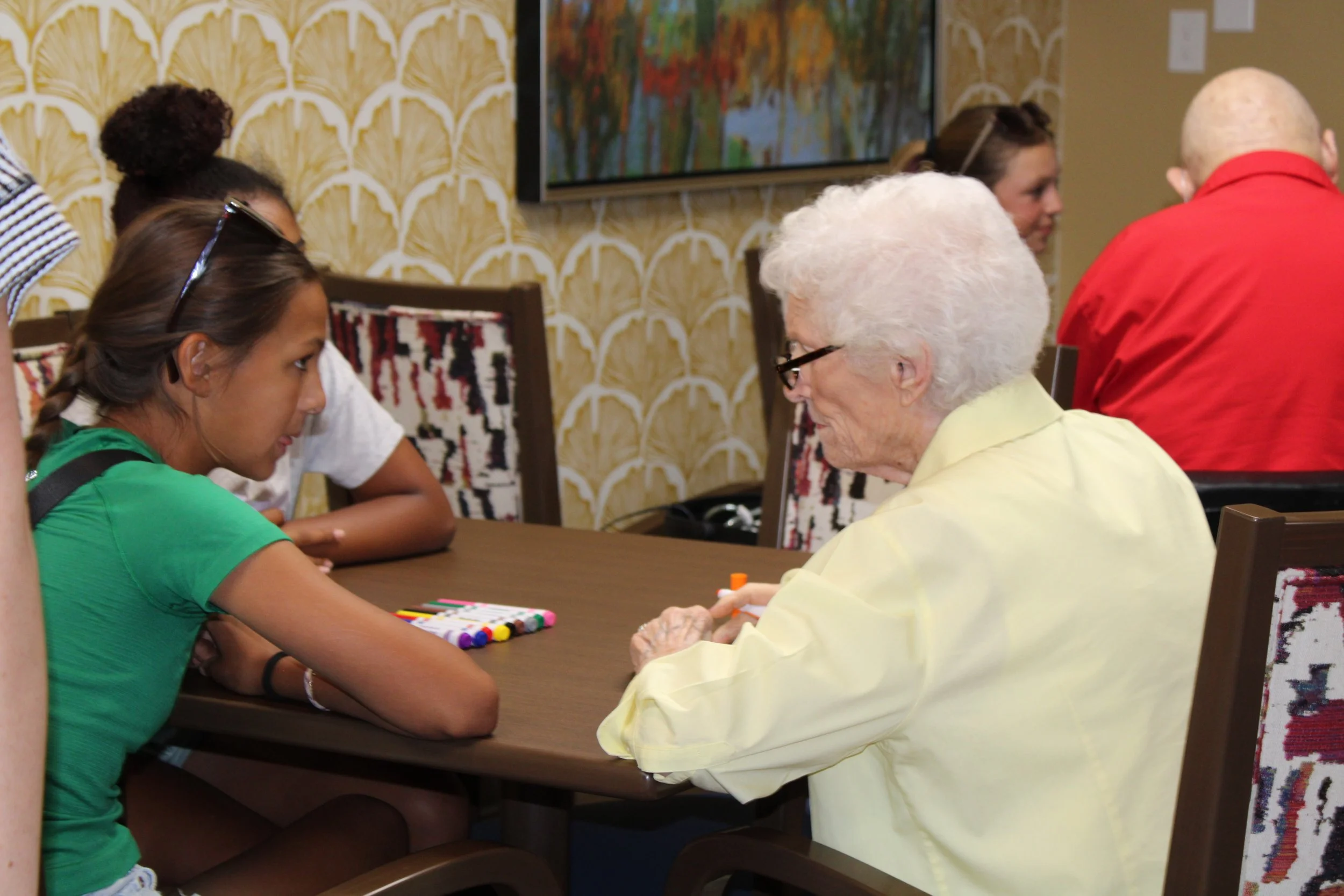 A young girl in a green shirt sitting at a table across from a smiling elderly woman with white hair, wearing glasses and a light yellow shirt, in a room with patterned wallpaper, engaged in a card game or activity.