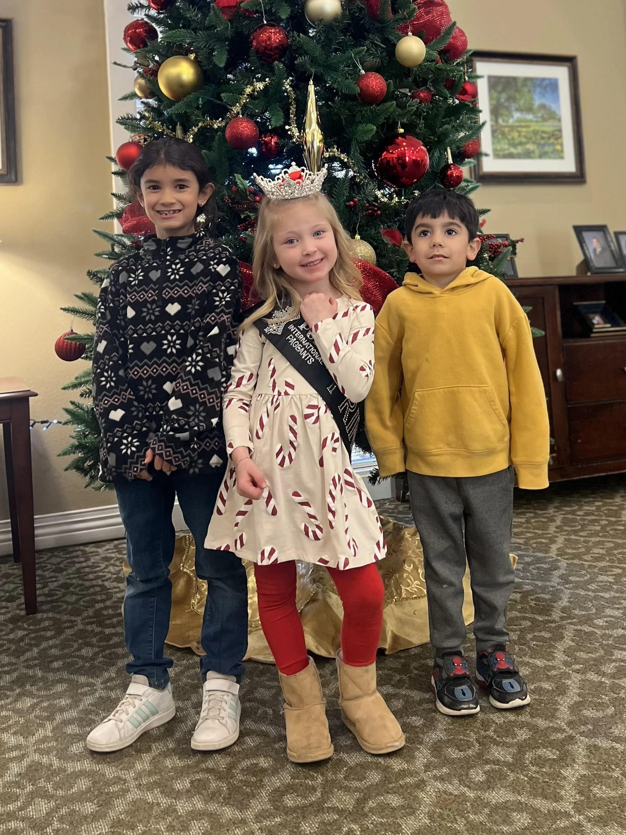 Three children stand in front of a decorated Christmas tree, smiling. The girl in the center wears a crown and a festive dress with candy cane patterns. The girl on the left wears a black sweater with snowflake and heart designs and white sneakers. The boy on the right wears a yellow hoodie and Spider-Man sneakers. The Christmas tree is adorned with red, gold, and black ornaments and gold garlands.