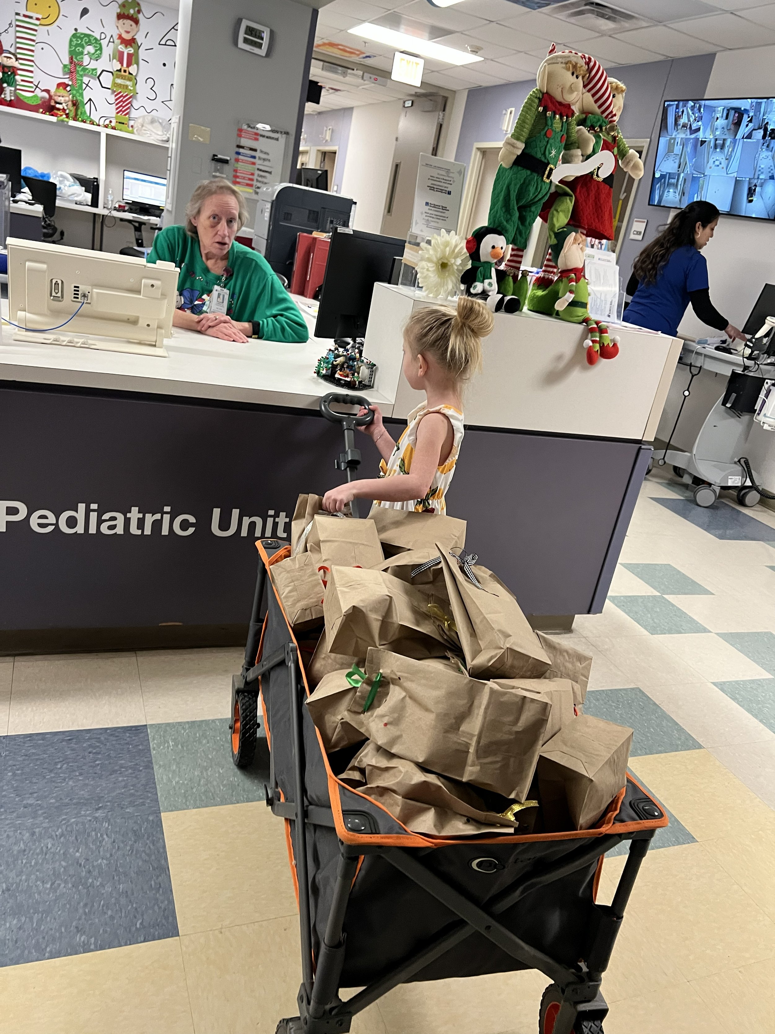 Child with blonde hair tied in a bun standing near shopping cart filled with paper bags in hospital lobby. Hospital staff and Christmas decorations in the background.