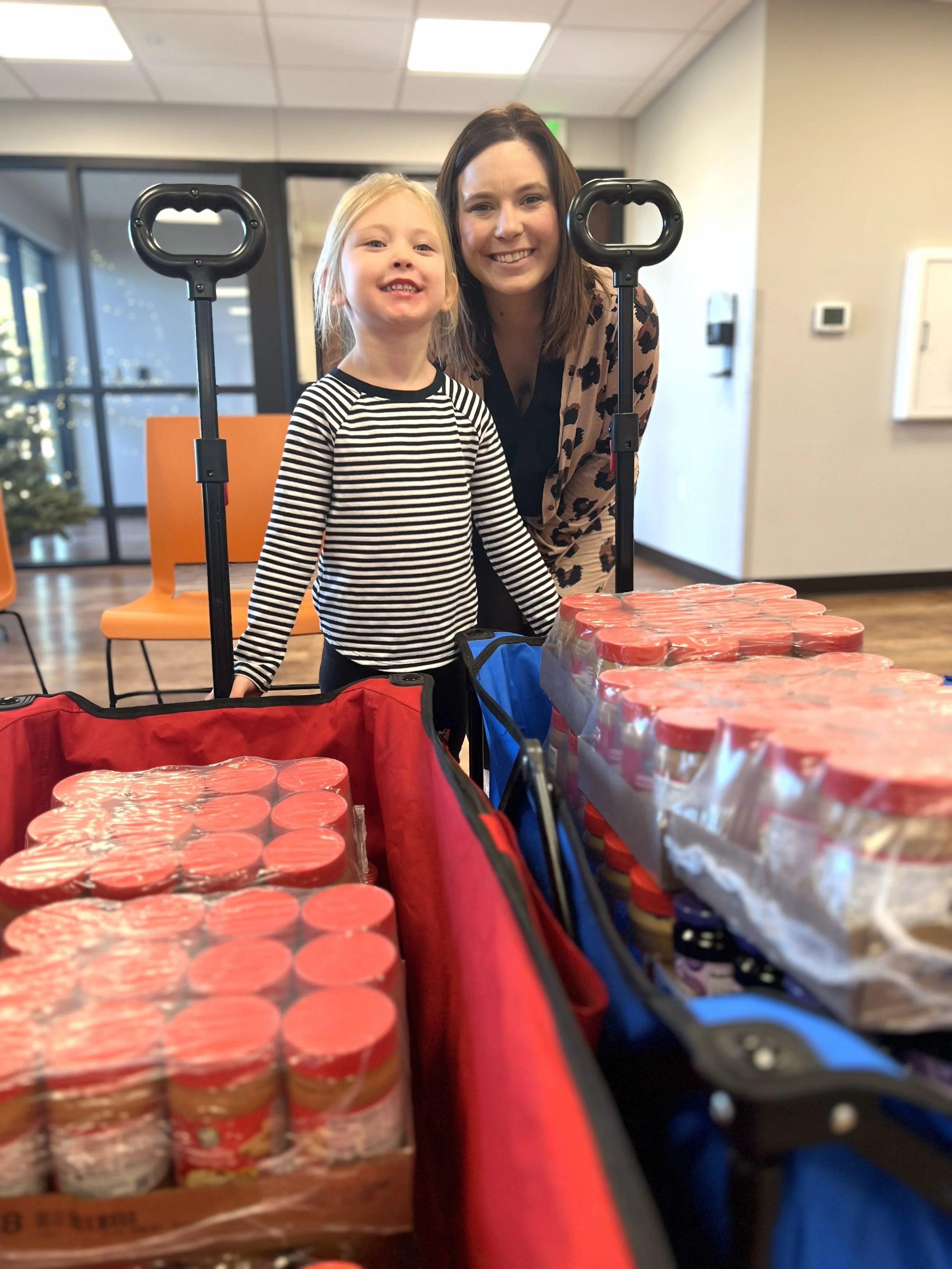 A smiling woman and a young girl are standing behind grocery carts filled with cases of canned goods in a store.
