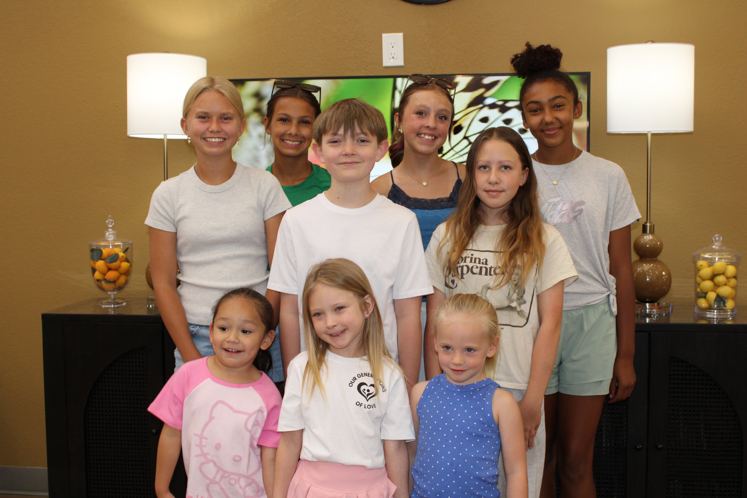 Group of young children and teenagers standing together indoors, smiling at the camera, with a TV screen and lamps in the background.