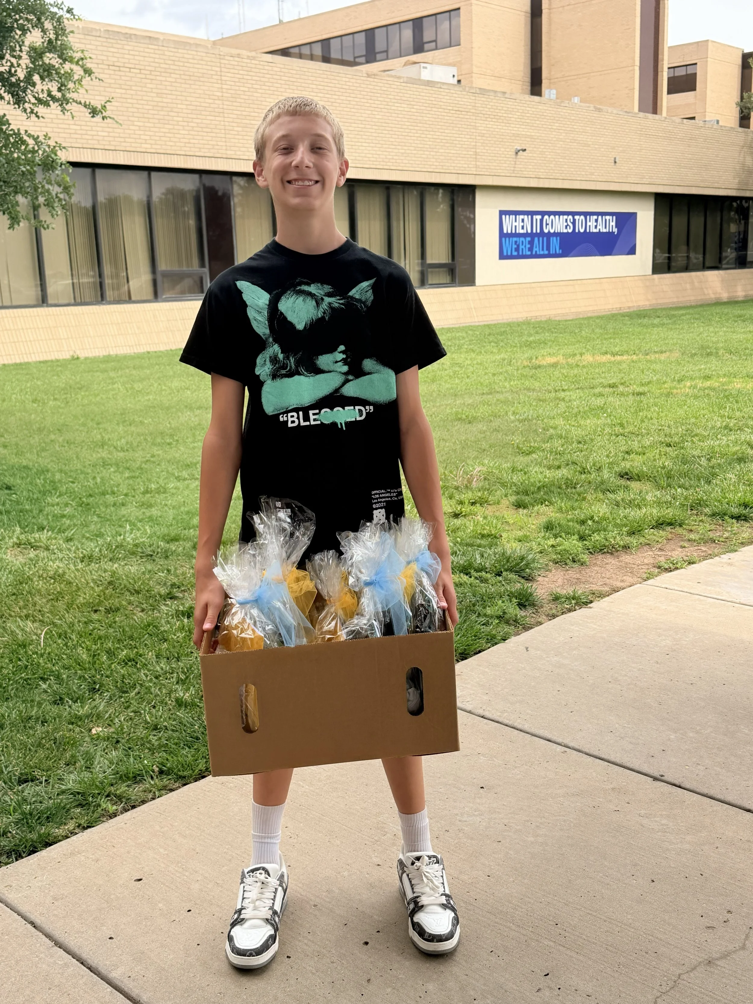 A boy standing outside on a sidewalk in front of a grassy area and a building, holding a cardboard box filled with clear plastic bags tied with blue and yellow ribbons.