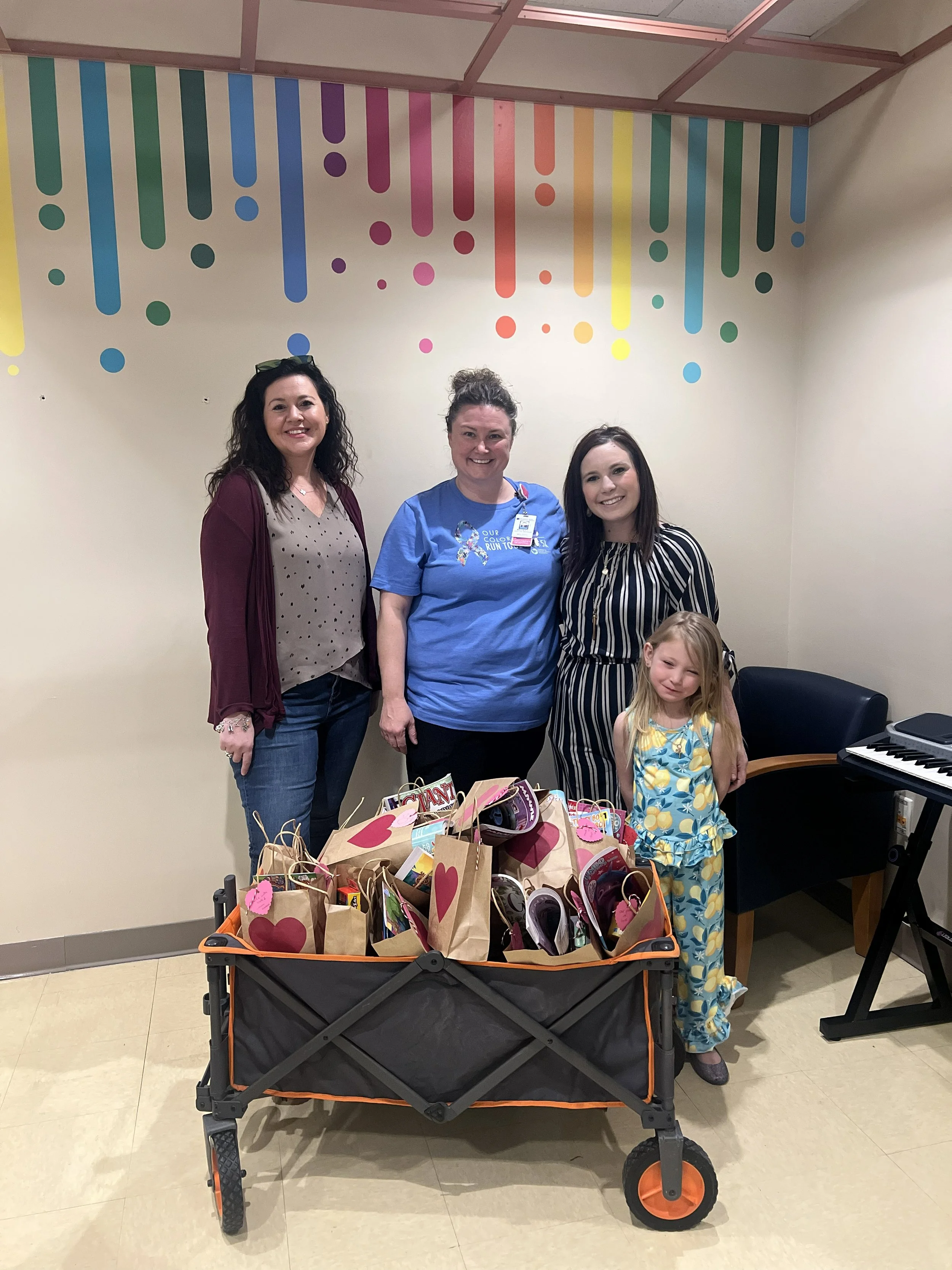 Four women and a young girl standing behind a shopping cart filled with gift bags decorated with hearts. The women are smiling, and the girl is standing in front of the cart. There is a colorful, abstract wall design behind them and a black electronic keyboard on the right side of the image.