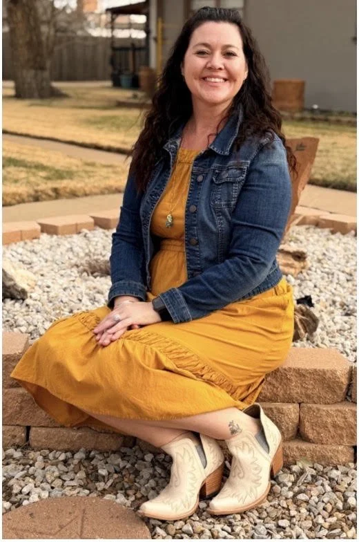 A woman sitting cross-legged outdoors on a stone border, smiling at the camera, wearing a mustard yellow dress, a denim jacket, and white cowboy boots.