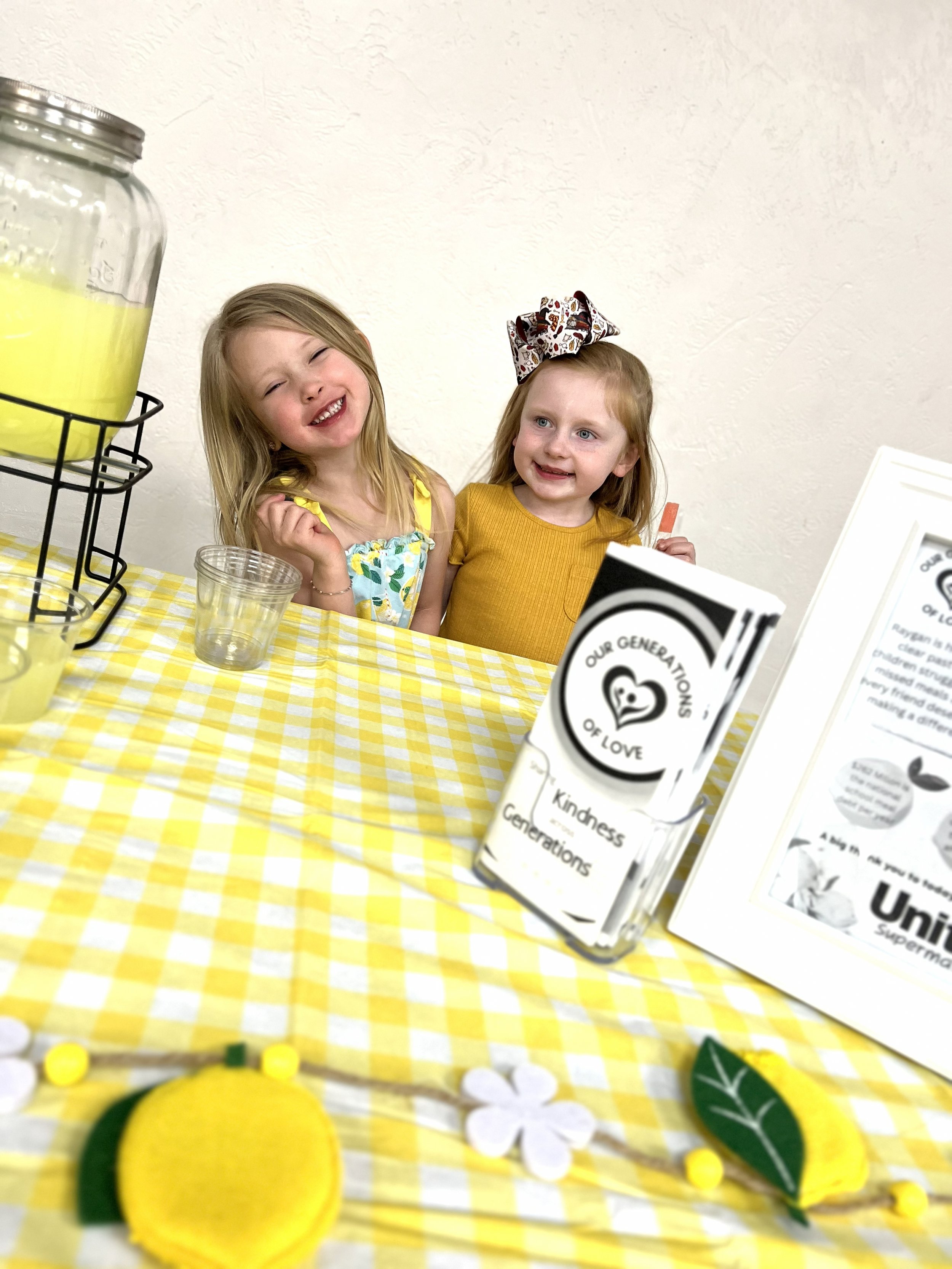 Two young girls smiling at a table with yellow checkered tablecloth, various items including a yellow felt lemon, paper flowers, and informational flyers about kindness and love.