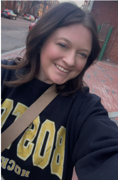 A young woman with brown hair smiling and taking a selfie outdoors on a brick sidewalk.