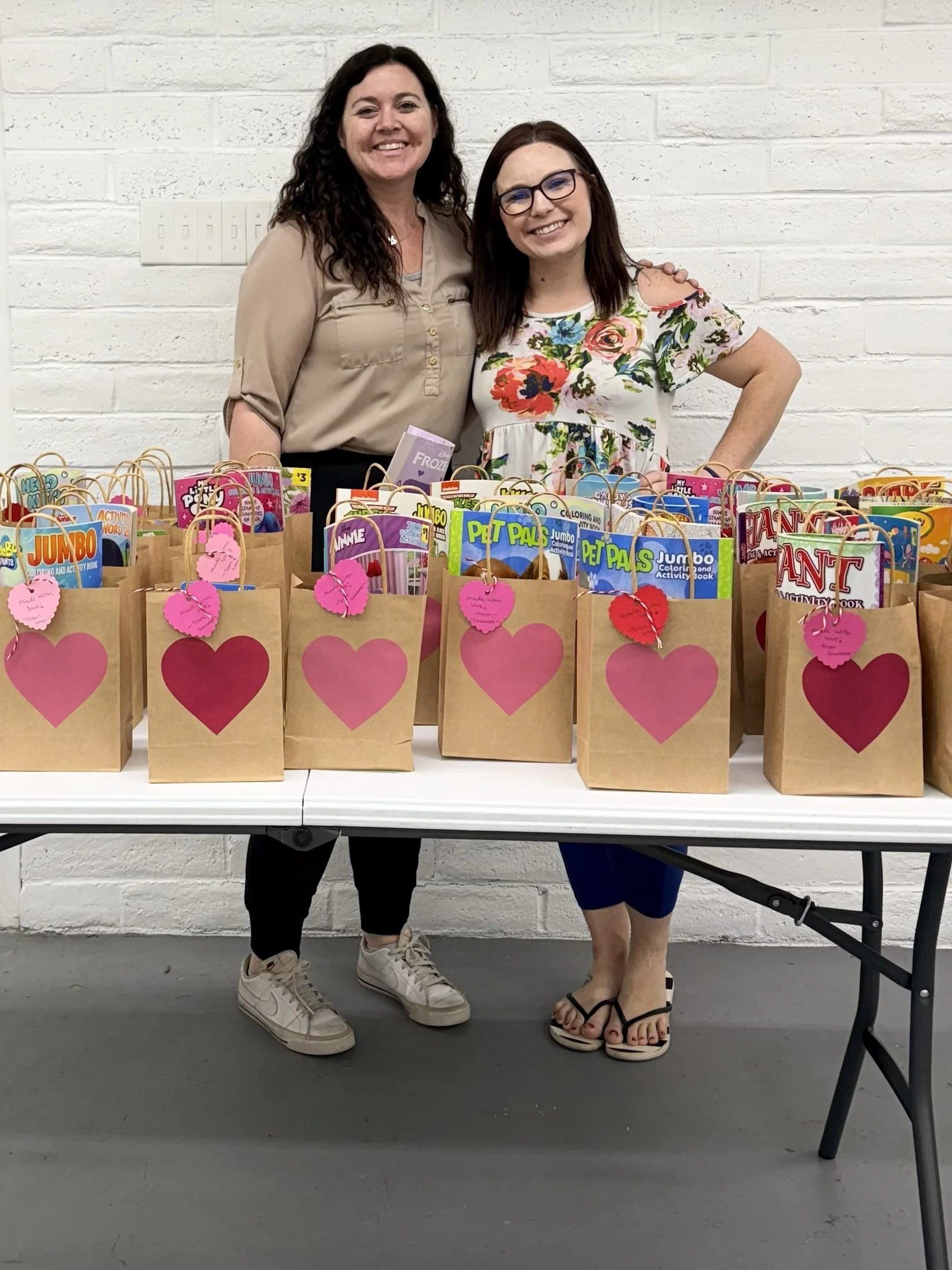 Two women standing behind a table filled with paper gift bags decorated with large pink hearts and tags, smiling at the camera against a white brick wall.