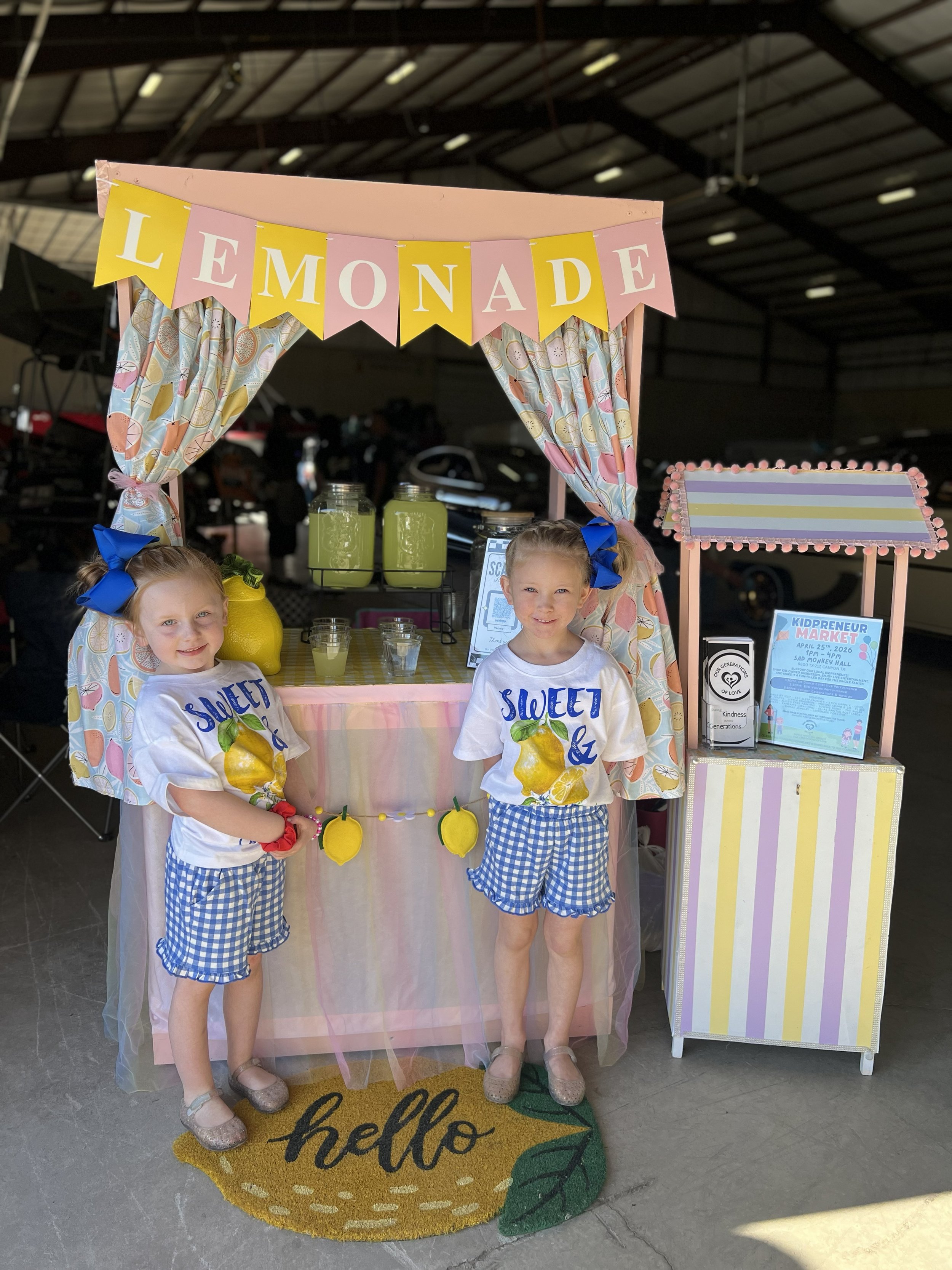 Two young girls with blue bows in their hair standing in front of a lemonade stand. The stand is decorated with a pink banner spelling 'LEMONADE' in alternating yellow and pink banners. The stand features jars of lemonade, a yellow lemon-shaped decoration, and signs including one for a Kidpreneur Market event.