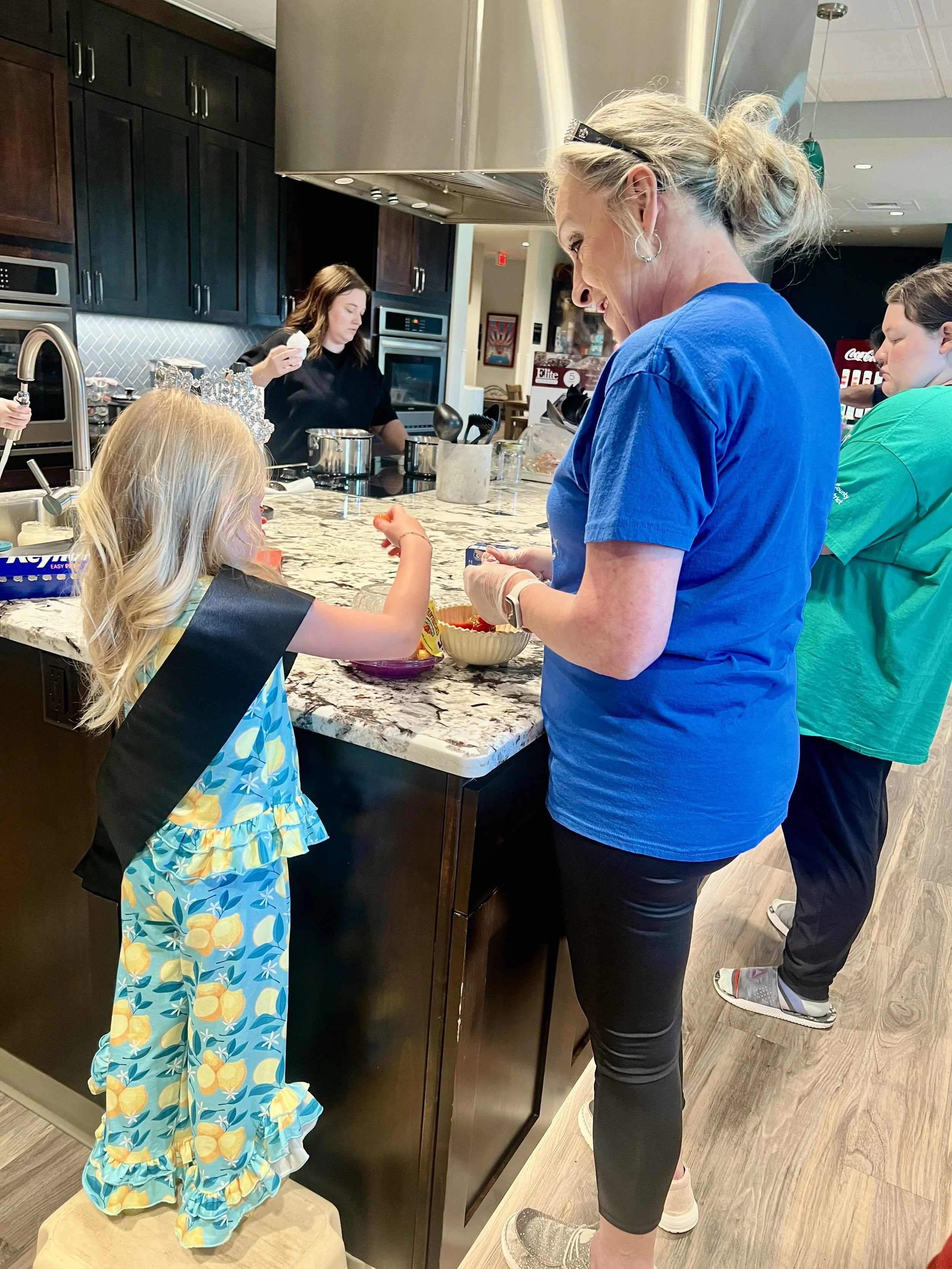 A woman in a blue shirt and black leggings prepares food at a kitchen counter, with a young girl in a colorful dress and apron standing on a stool nearby, and two other women in the background.