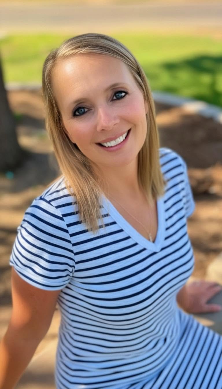 A woman with blonde hair and blue eyes smiling at the camera, sitting outdoors on a sunny day, wearing a white and navy striped shirt.