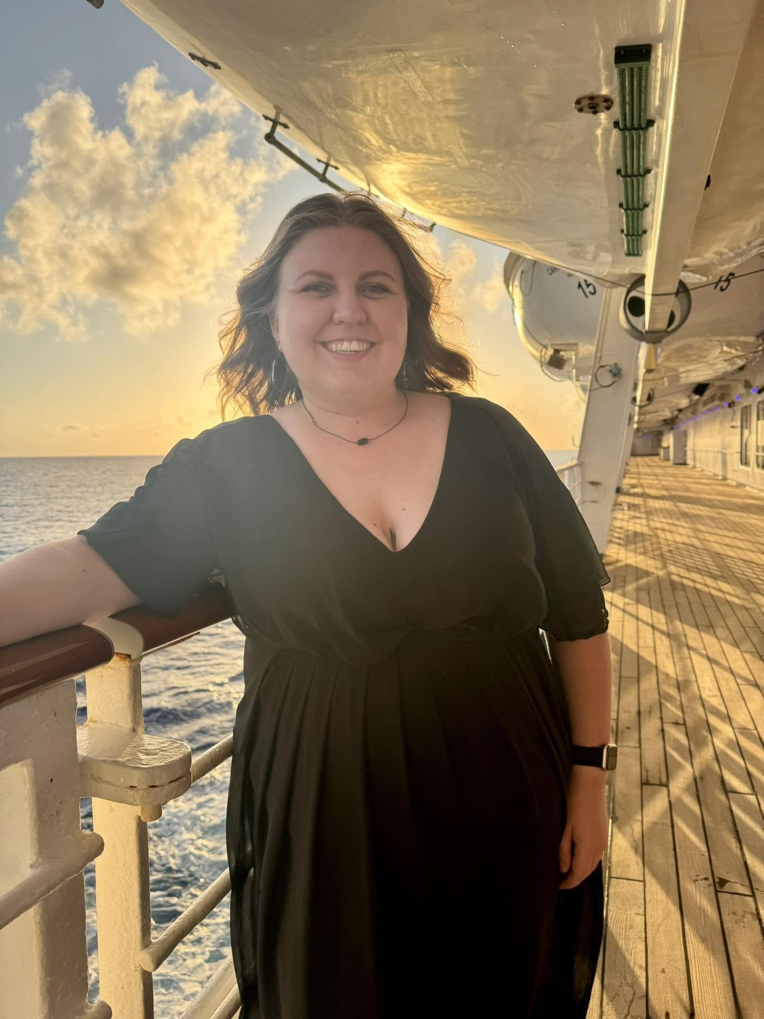 A woman with shoulder-length wavy hair smiling, wearing a black dress and a necklace, standing on a ship's deck with a sunset sky and ocean in the background.