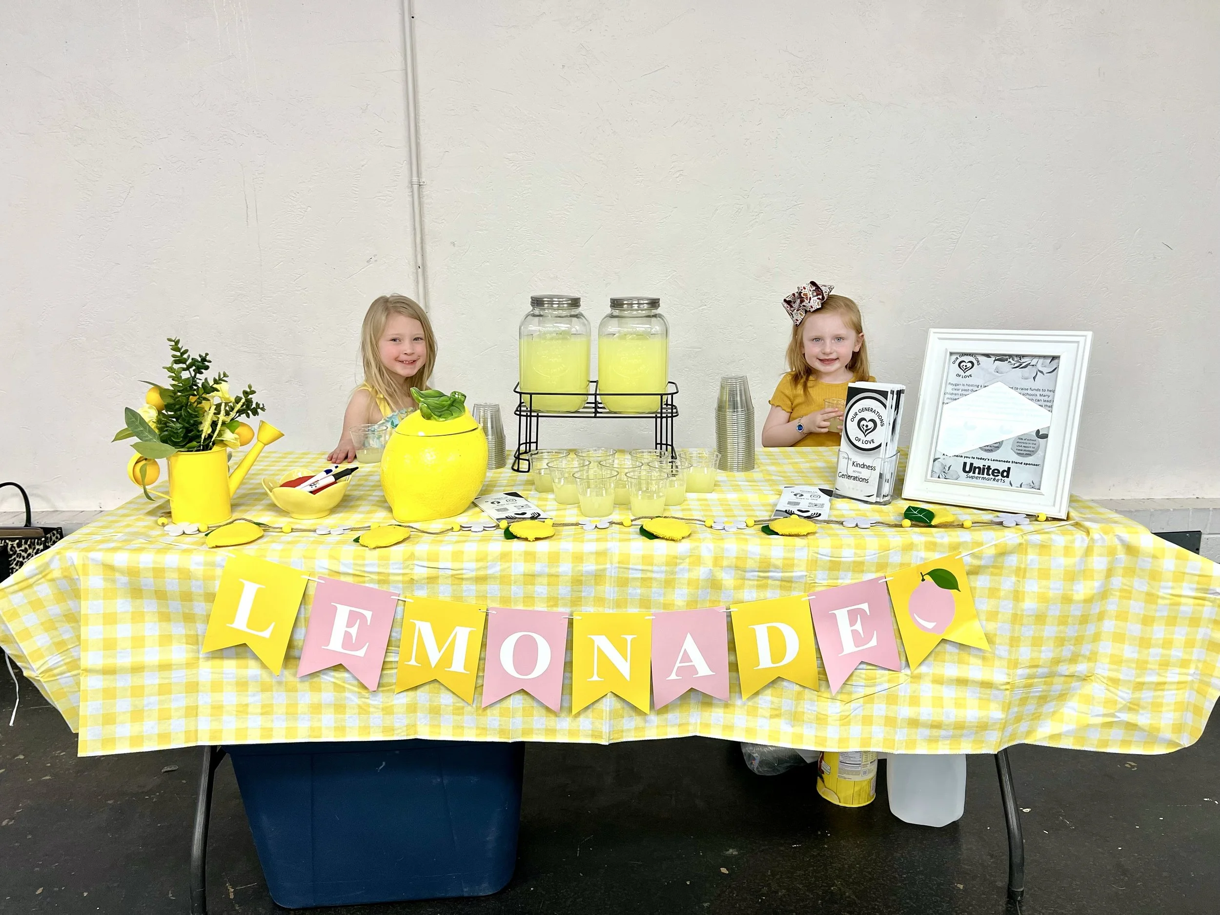 Table with lemon-themed decorations, including a yellow watering can, a lemon-shaped container, a plant, and a lemonade stand with cups and jars of lemonade. Two young girls sit behind the table, smiling. A framed sign and a colorful banner that reads 'Welcome' are also visible.