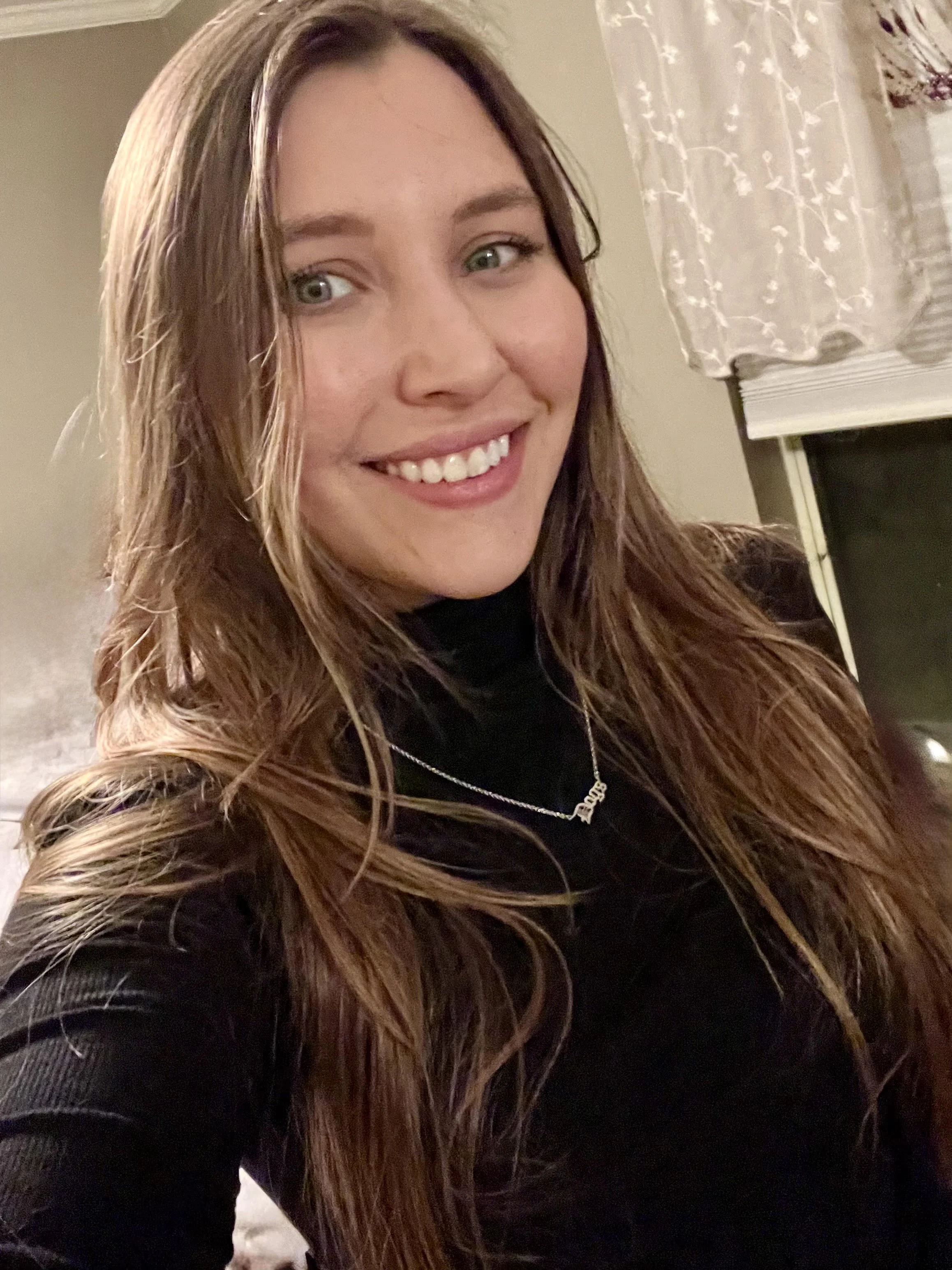 A smiling woman with long, wavy, brown hair and green eyes, wearing a black top and a necklace, indoors near a window with a patterned curtain.
