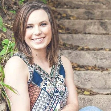 A young woman with shoulder-length brown hair, smiling, wearing a patterned sleeveless top, sitting outdoors near a wooden staircase surrounded by greenery.