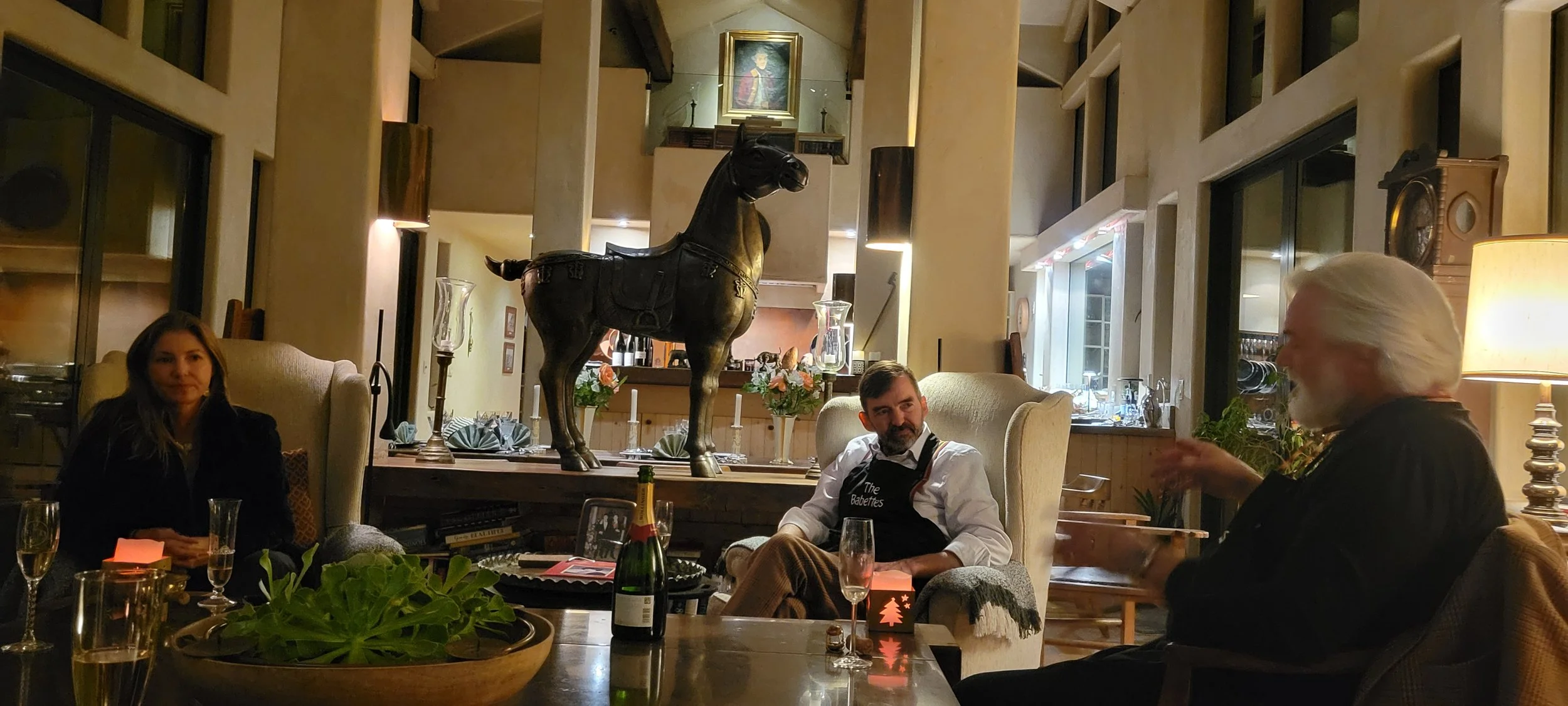 A cozy restaurant or cafe setting with three people sitting around a table, decorated with flowers, candles, and champagne glasses. A large black horse statue stands on a table behind them, with a royal portrait hanging on the wall above.