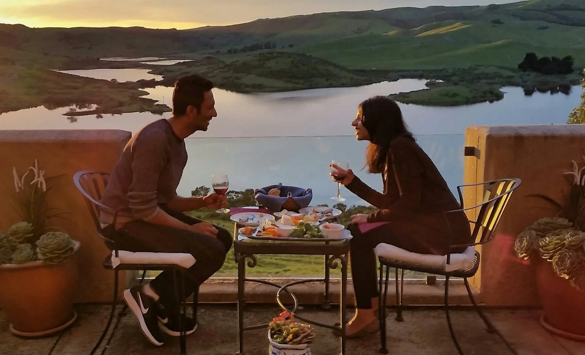 A man and woman enjoying a meal together on an outdoor terrace overlooking a river and green hills at sunset, with wine glasses and a cheese platter on the table.