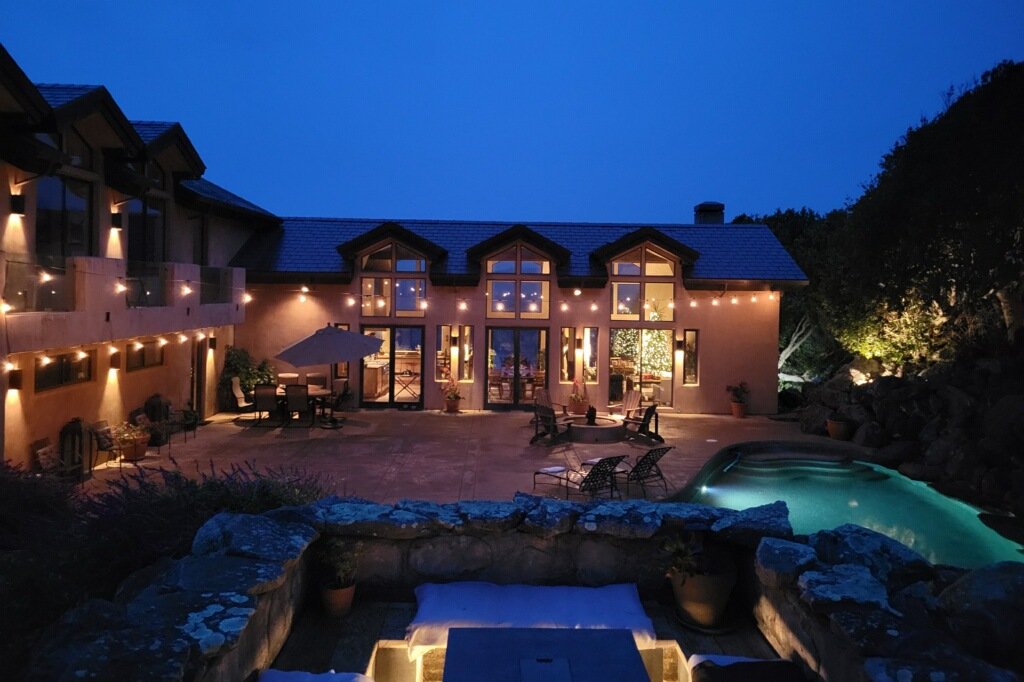 A beautiful house at night with warm lights, a backyard patio with furniture, an umbrella, string lights, and a small pool or hot tub, surrounded by rocks and trees.