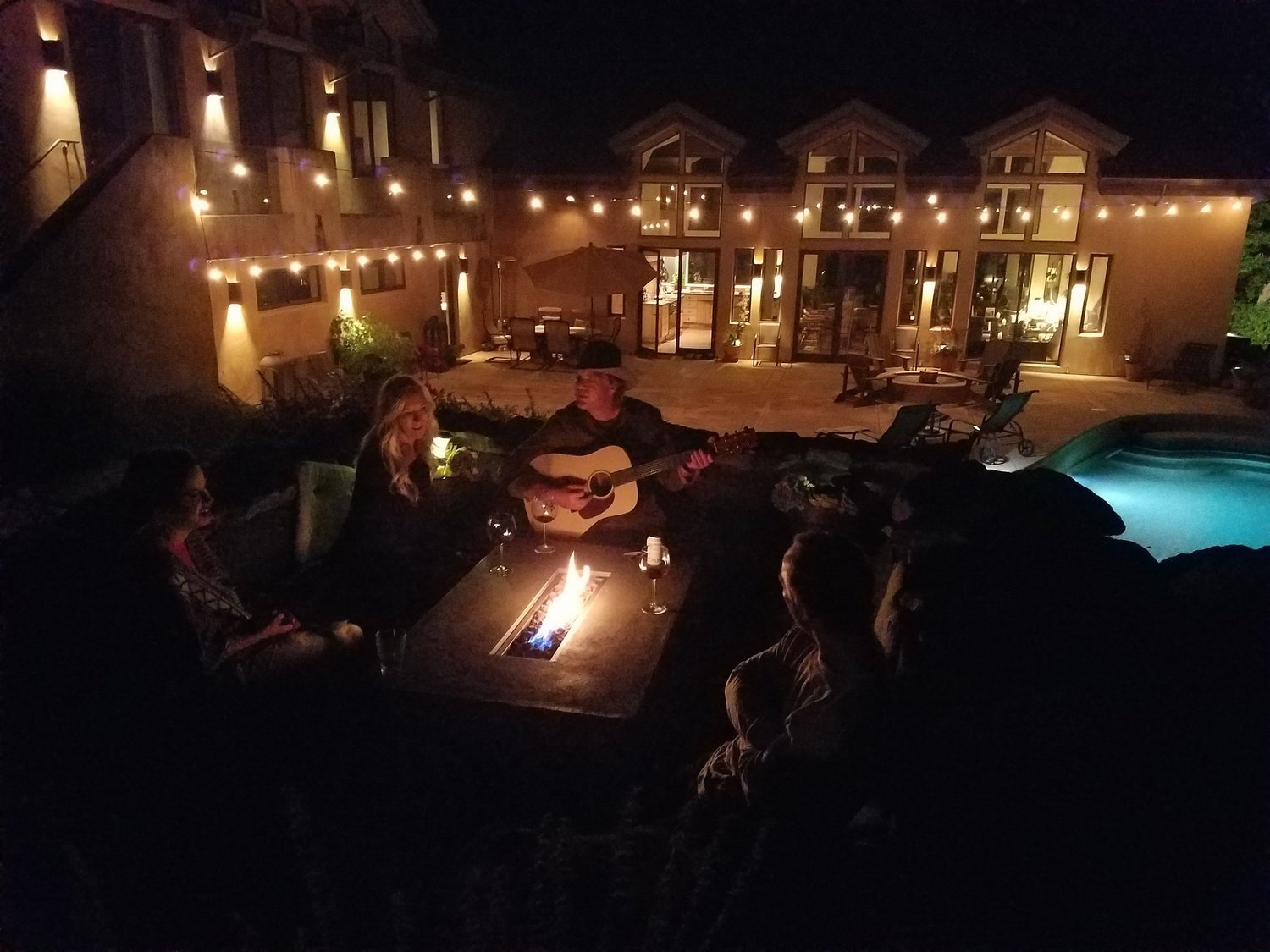 People enjoying a nighttime outdoor gathering by a fire pit with a guitarist and singer, patio furniture, string lights, and a swimming pool in the background.