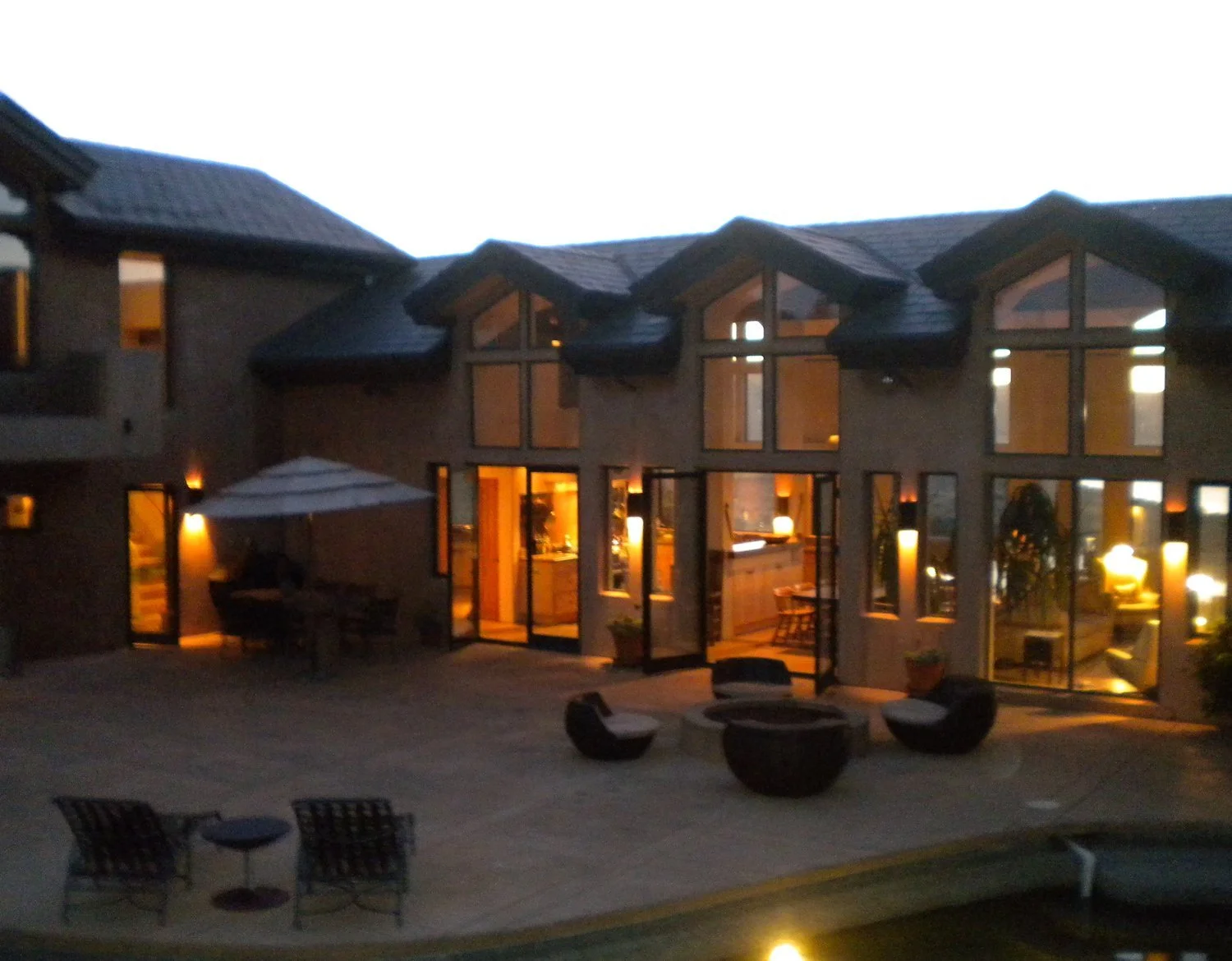 Night view of a modern house with large glass windows and doors, showing warm indoor lighting. Outdoor patio area with seating, a table with an umbrella, and decorative fire pits or planters.
